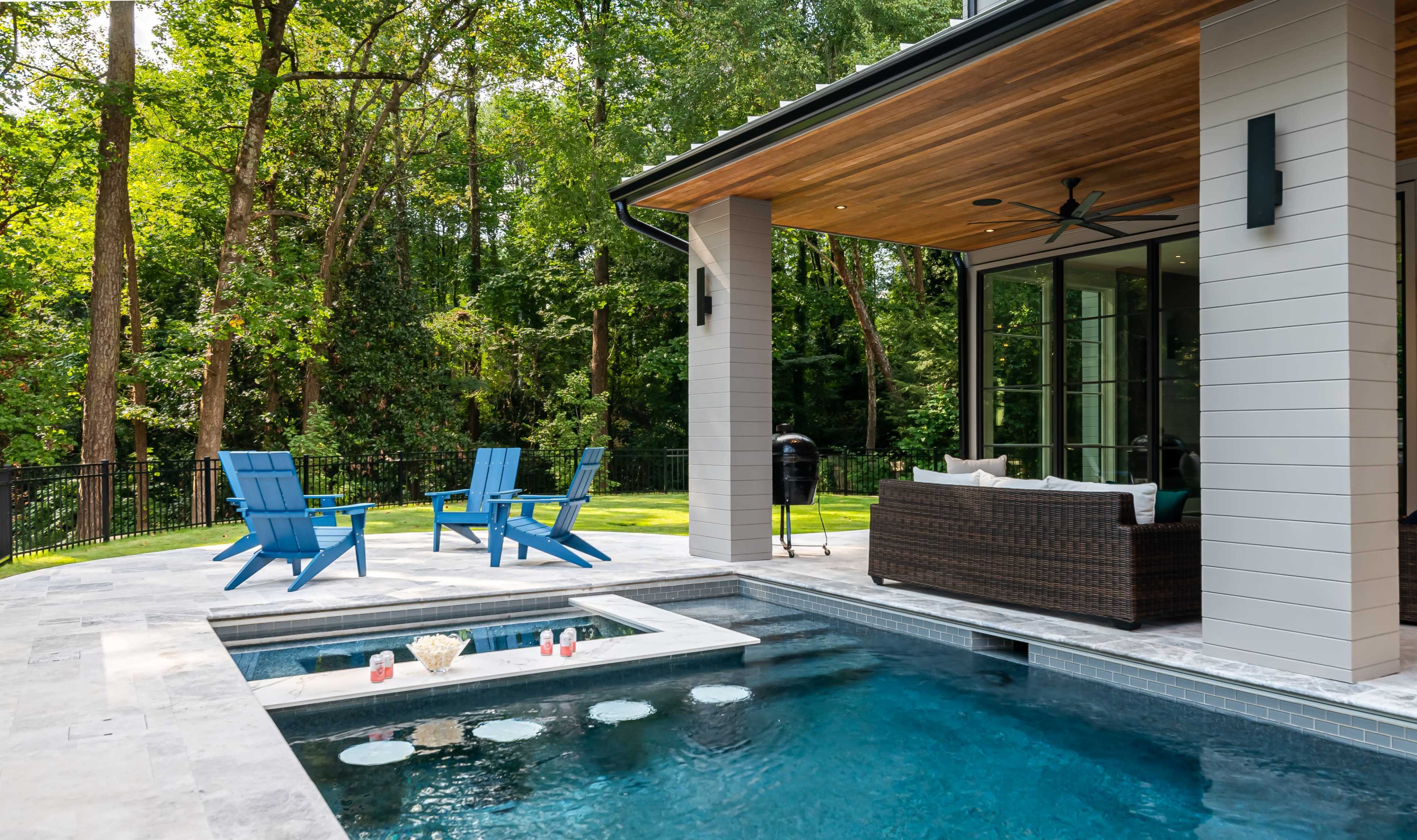 The scene features a modern outdoor patio with a swimming pool, lounge chairs, and a shaded seating area next to a wooded landscape.