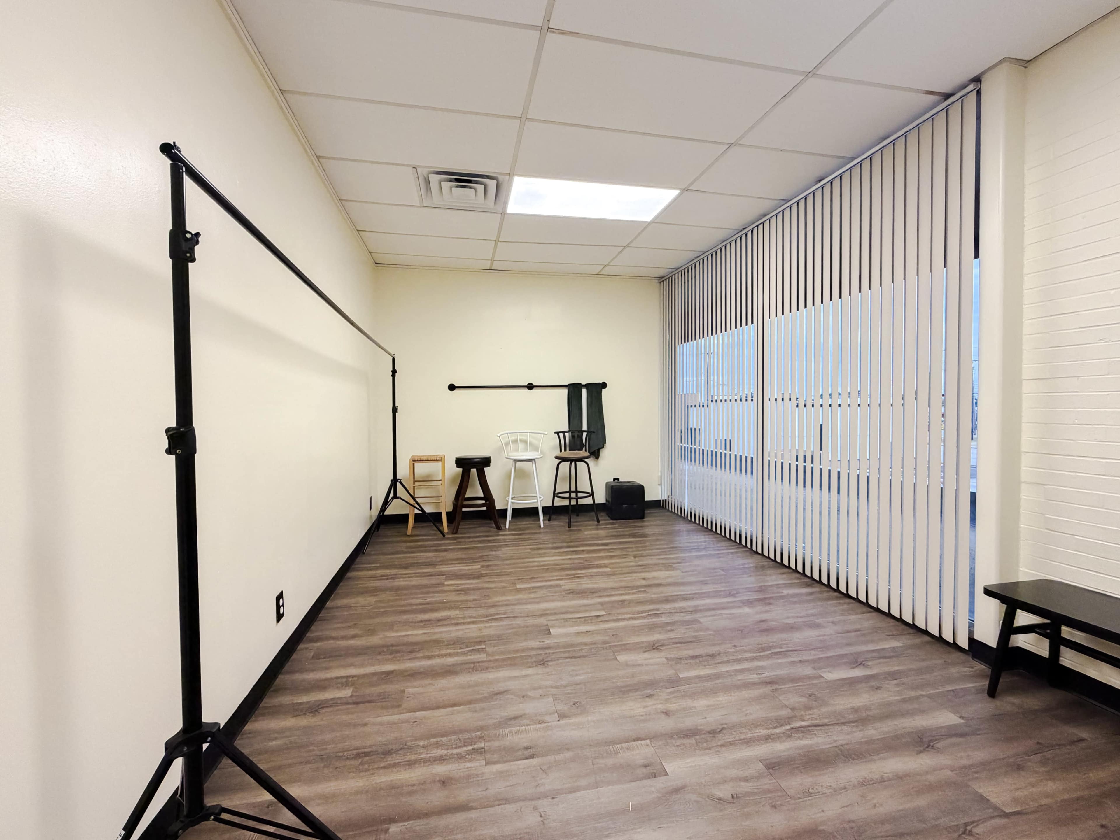 The image shows an empty room with wooden flooring, featuring a backdrop stand and various chairs against the wall, as well as vertical blinds covering a large window.