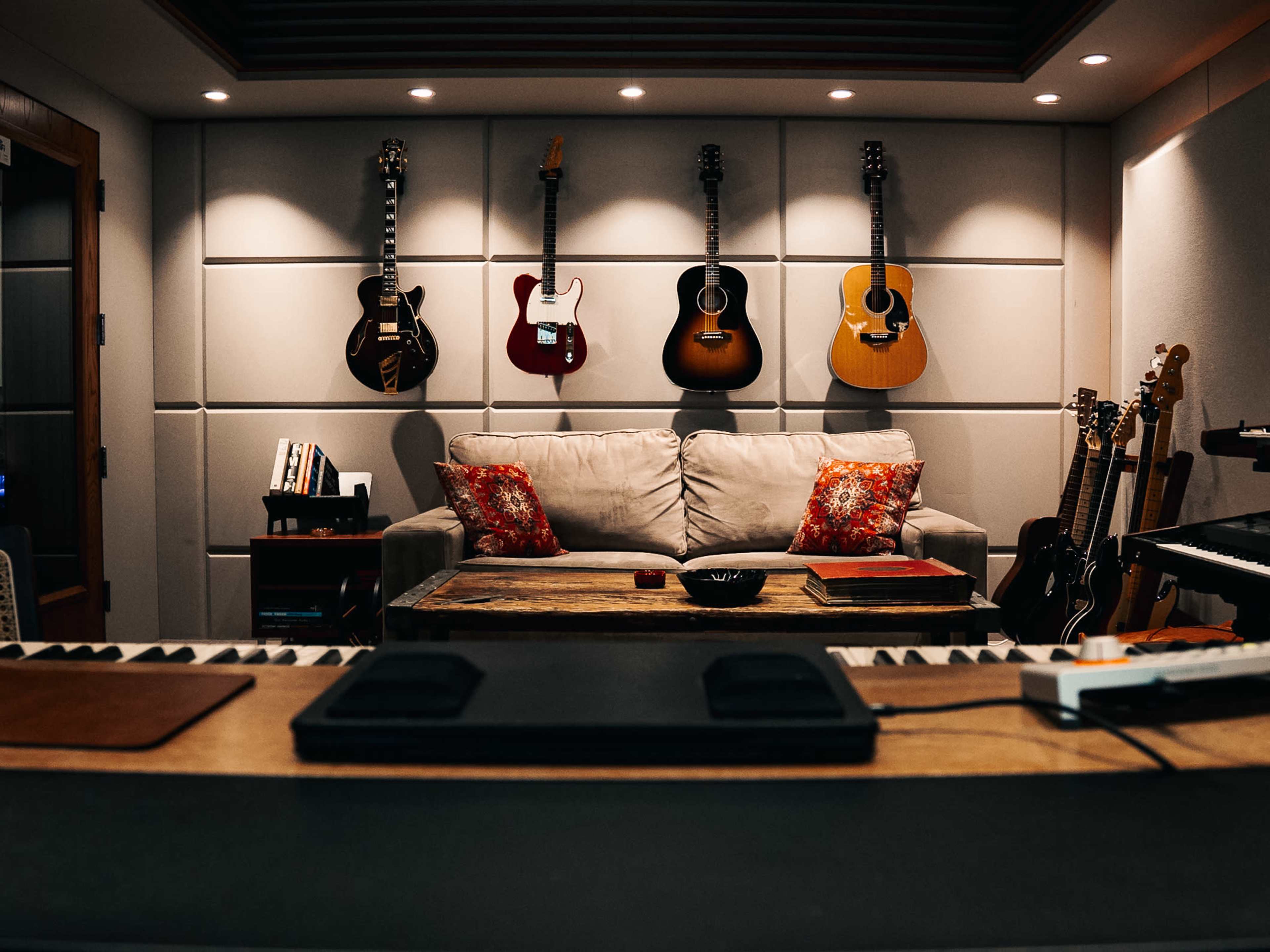 A cozy music studio features a sofa with decorative pillows, surrounded by various guitars hanging on the wall and a keyboard in the foreground.
