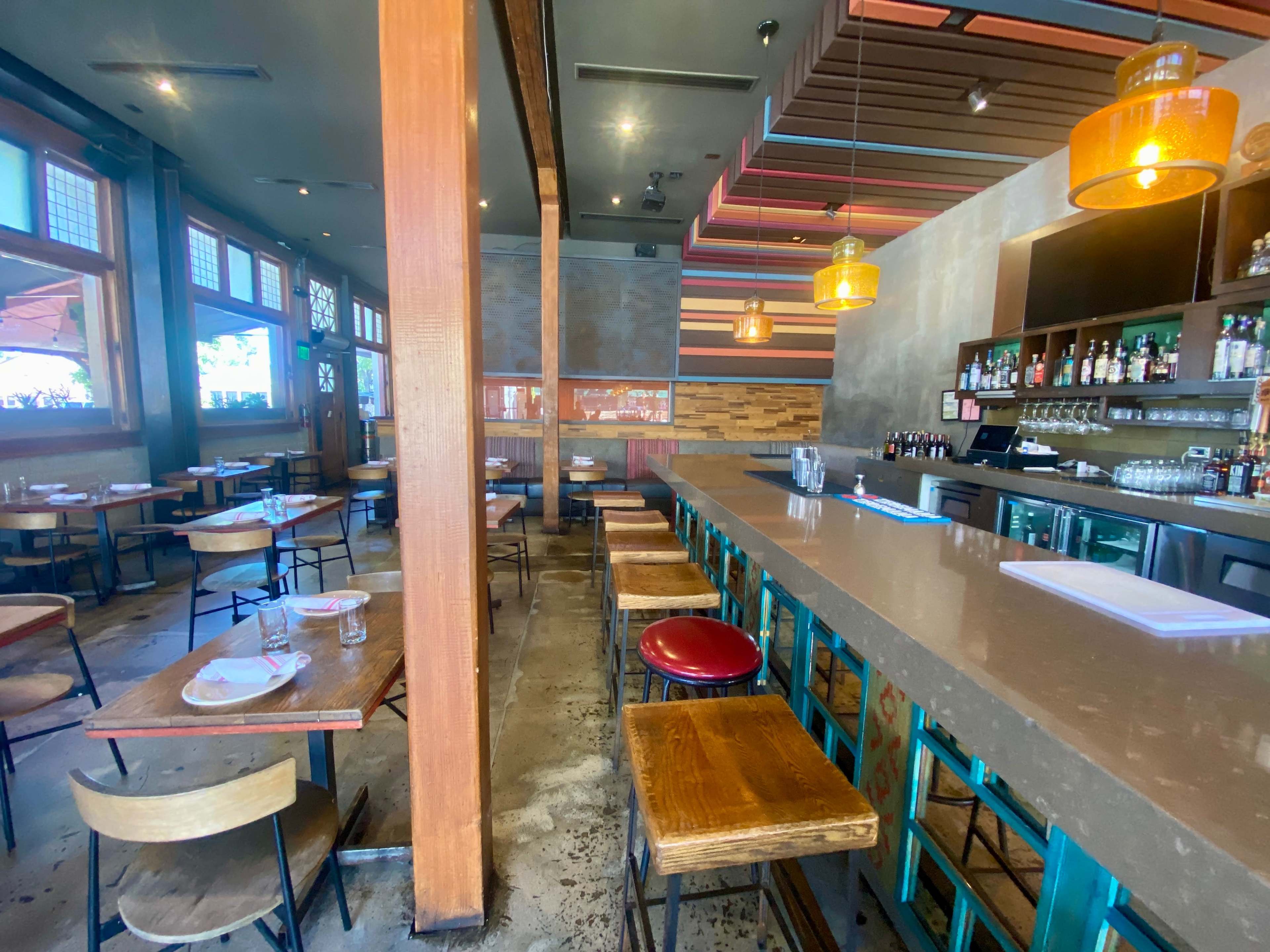 A restaurant interior featuring a long bar with stools, wooden tables, and large windows that provide natural light.