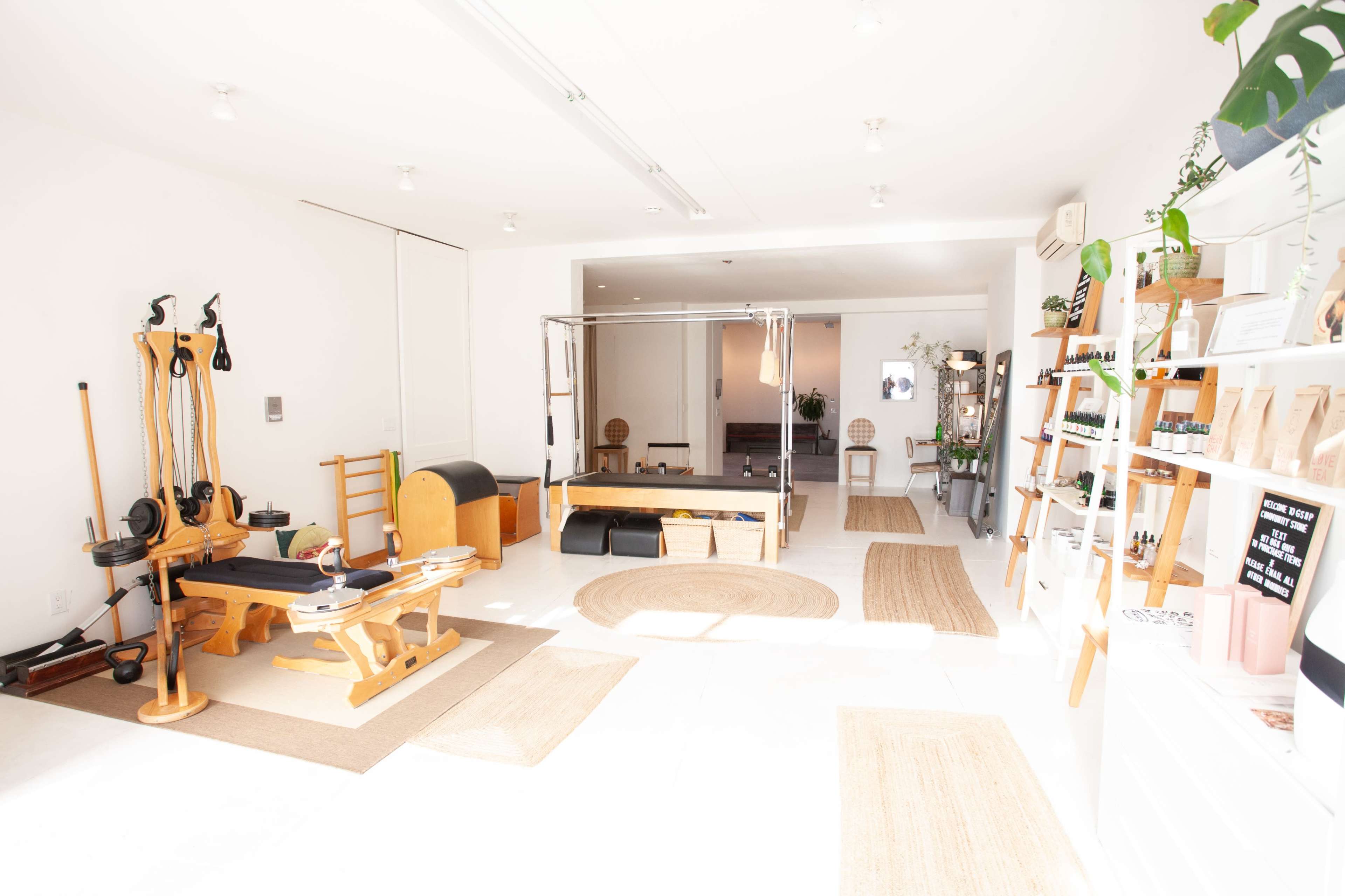 The image shows a bright, minimalist wellness studio featuring Pilates equipment, wooden furniture, and natural light illuminating the space.