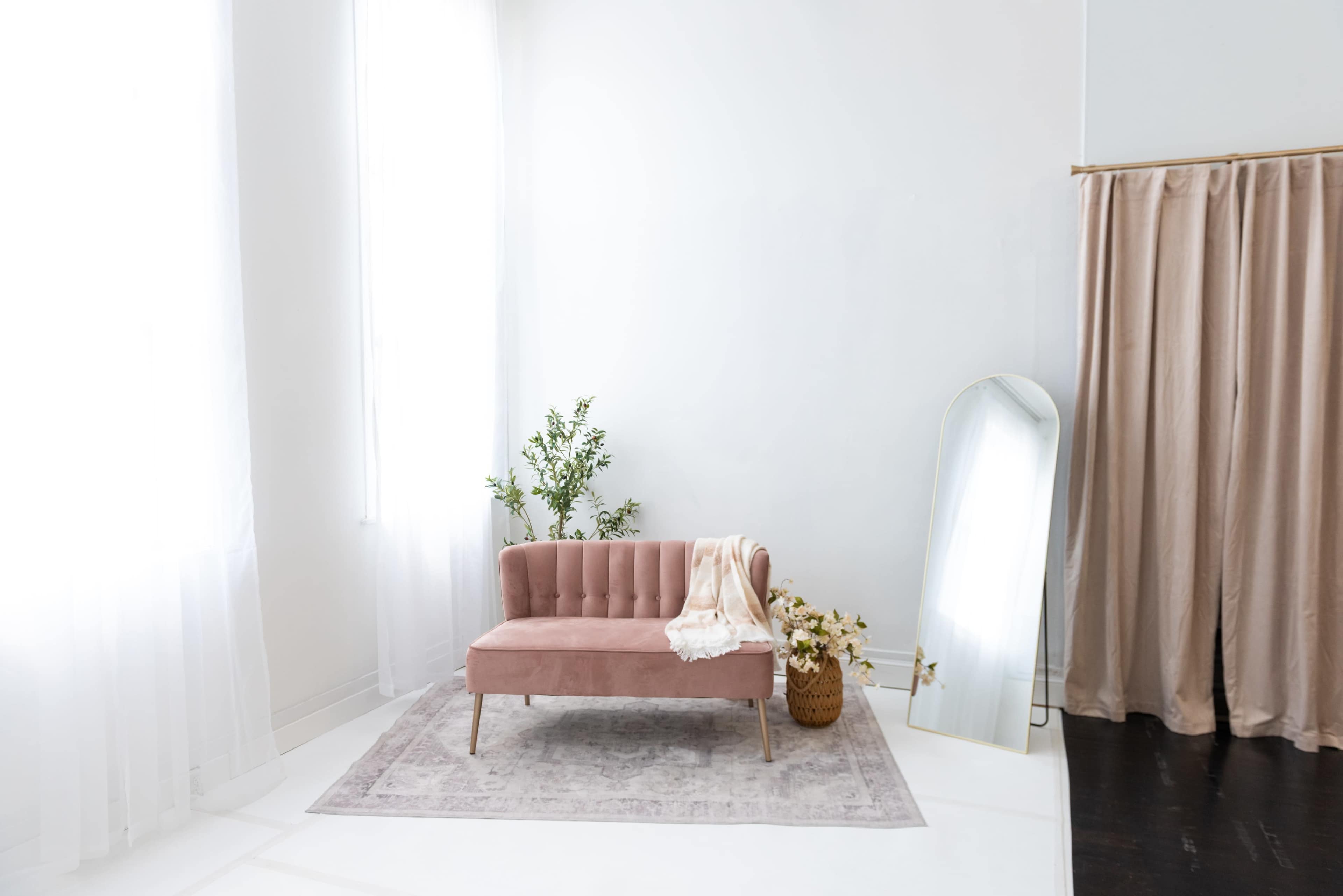 The image shows a minimalist living room with a pink velvet sofa, a decorative mirror, a potted plant, and sheer curtains allowing natural light to enter.