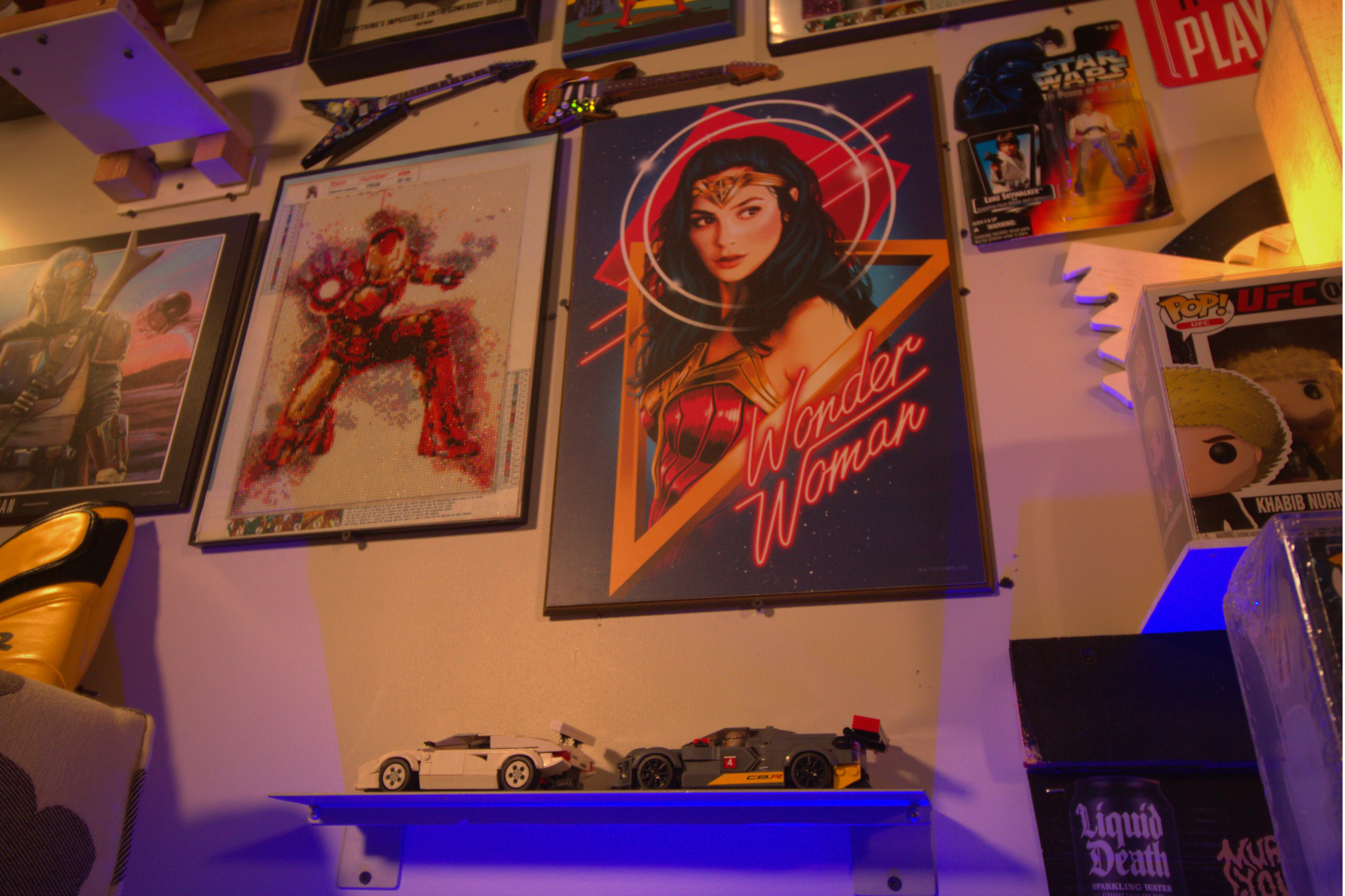 The scene features a wall adorned with framed posters of Iron Man and Wonder Woman, along with a display of miniature toy cars on a shelf below them.