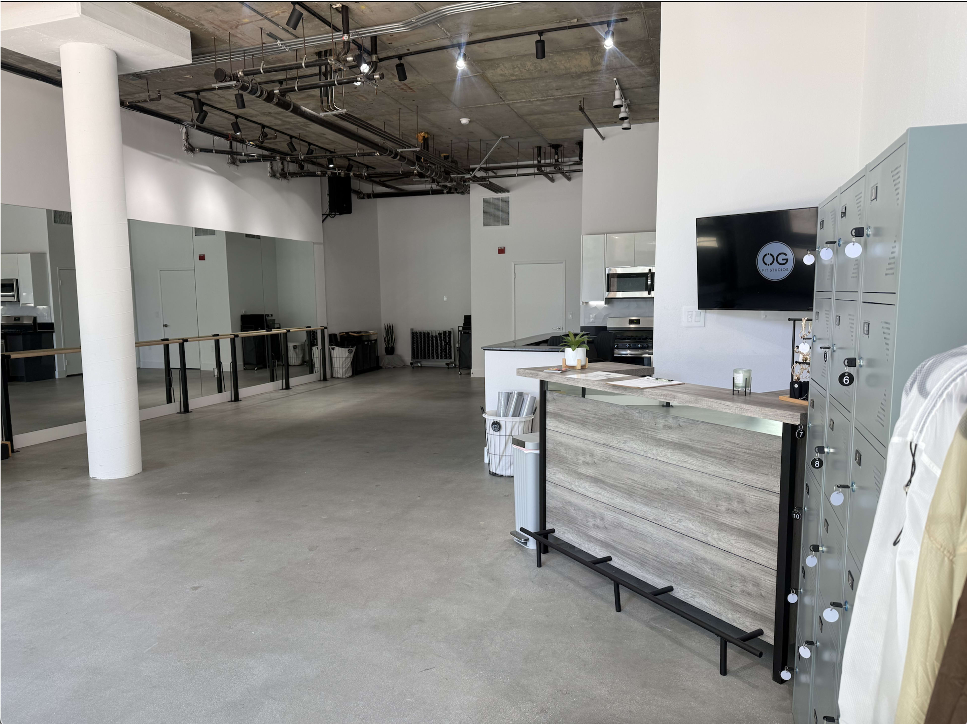 The image shows a spacious, minimalist interior that features a reception area with lockers, a bar-like counter, and mirrors along one wall in an open layout.