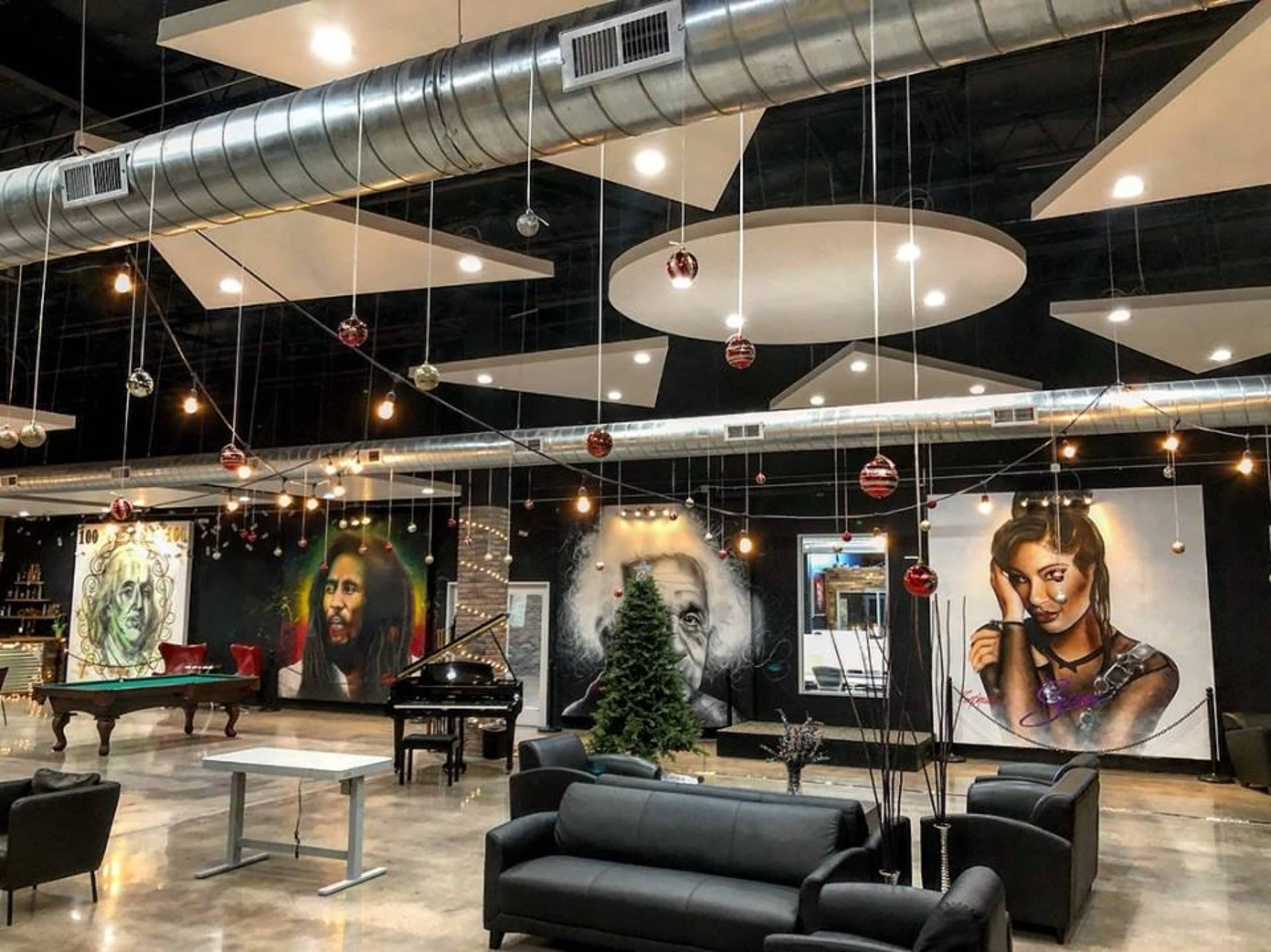 The interior of a modern studio space features a black sofa arrangement, a grand piano, and vibrant wall murals of famous figures, along with decorative lighting and a Christmas tree.