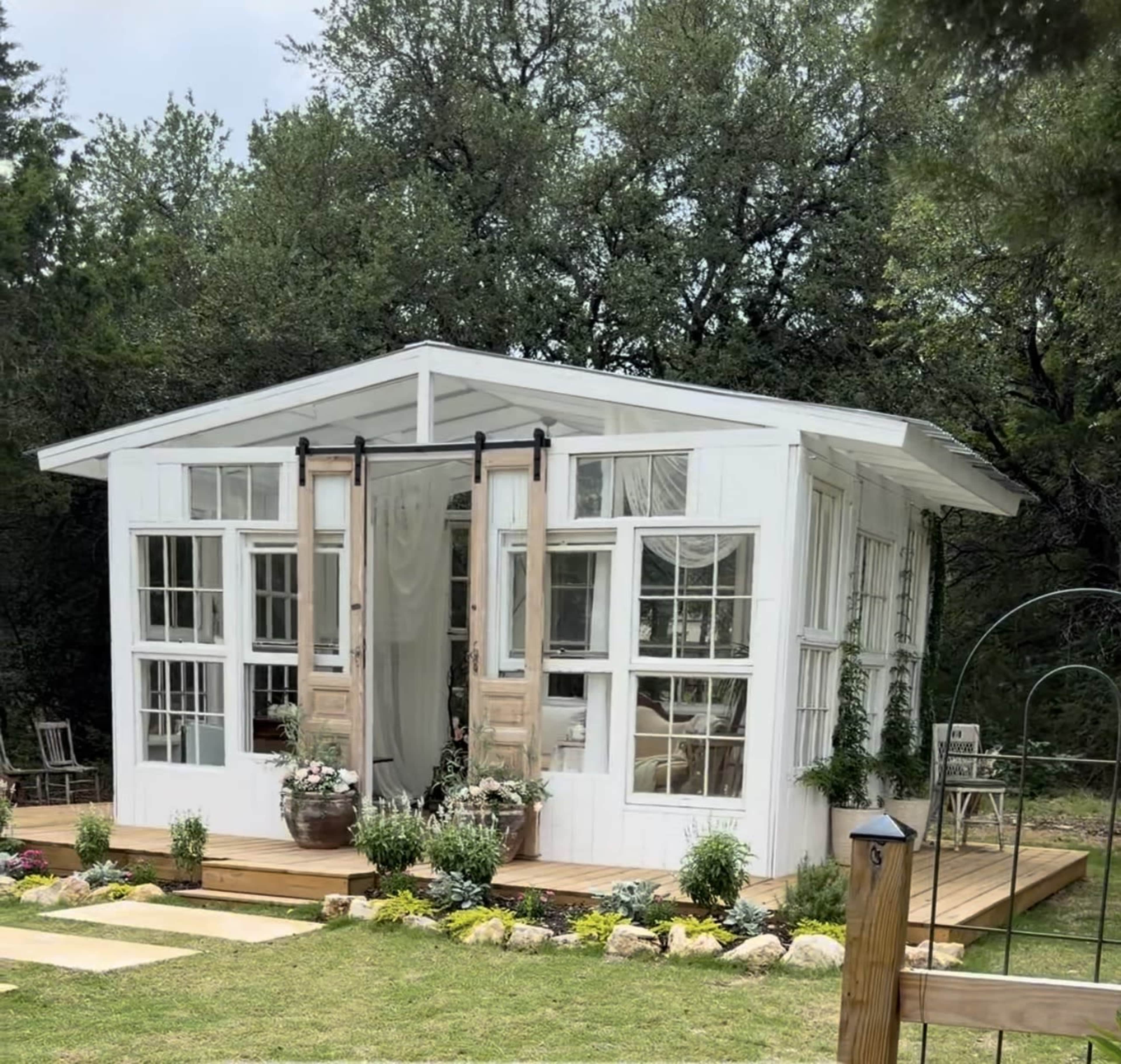 The image shows a small, white wooden building with multiple windows and a wooden deck surrounded by greenery and flowers.