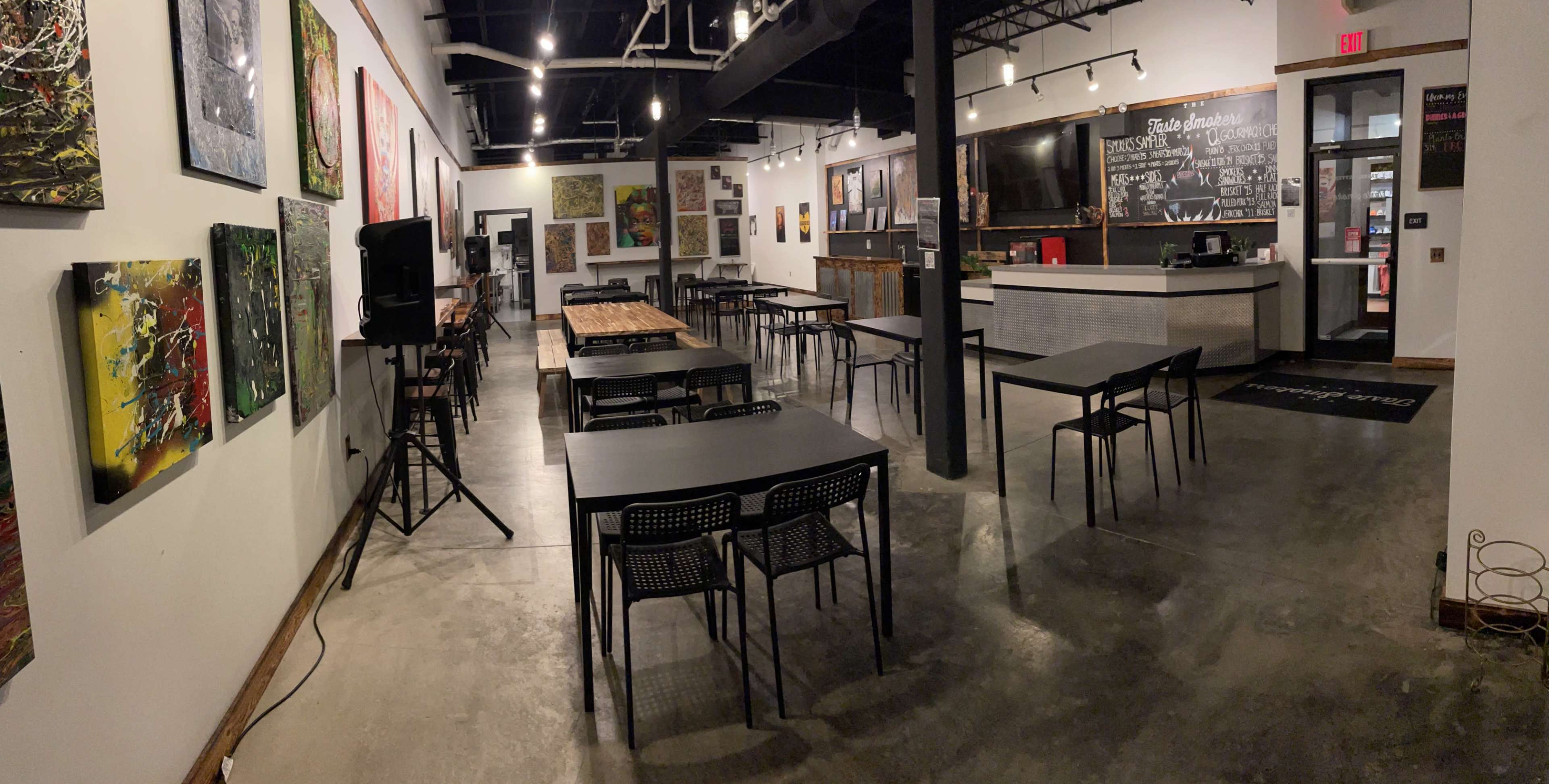 An indoor space with multiple tables and chairs arranged around an art-filled room, featuring a bar area in the background.