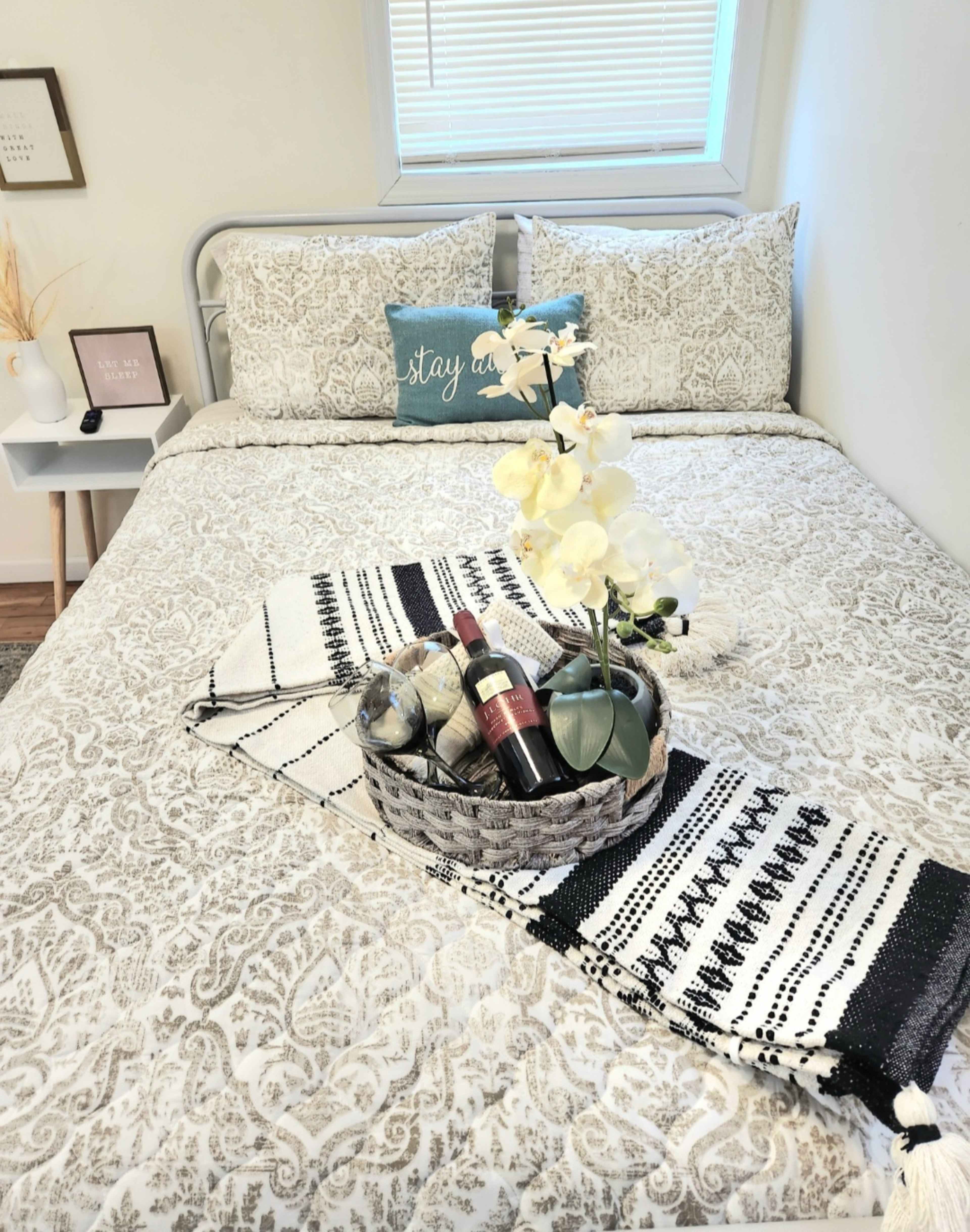 A neatly made bed with patterned bedding features a woven basket containing a bottle of wine and decorative items, placed on a decorative throw.