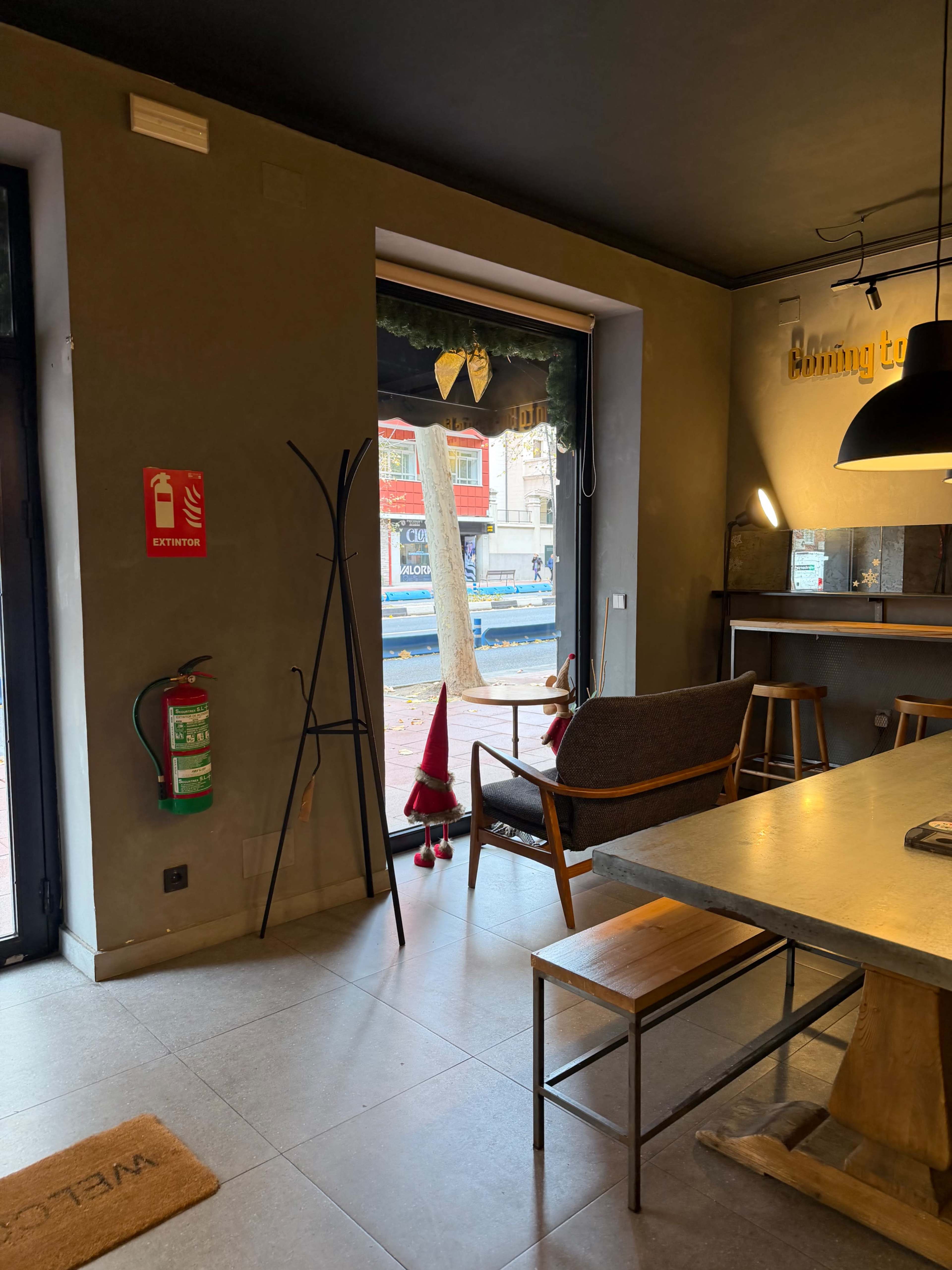 The image shows a modern café interior with a wooden table, chairs, and a large window displaying a street view outside, along with a small decorative gnome positioned near the entrance.