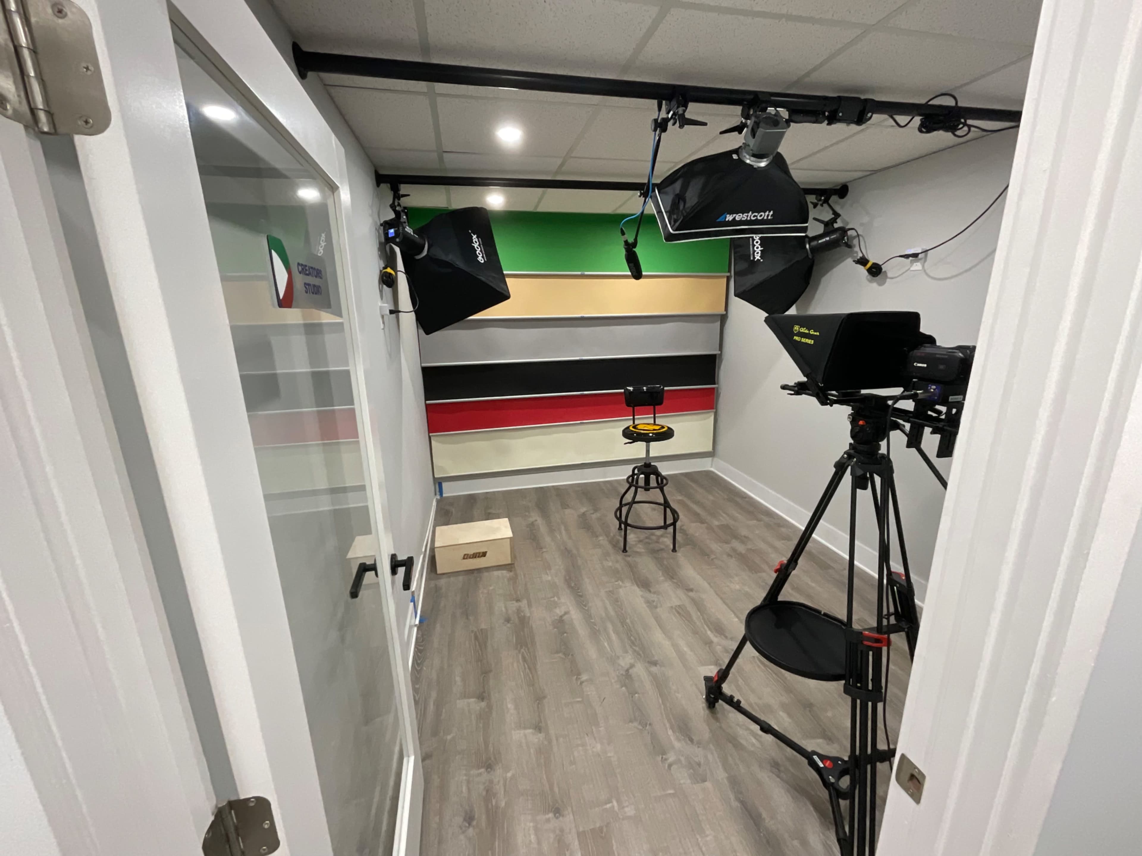 The image shows a small video production studio equipped with lighting kits and a camera set on a tripod, featuring a colorful striped backdrop and a stool in the center.