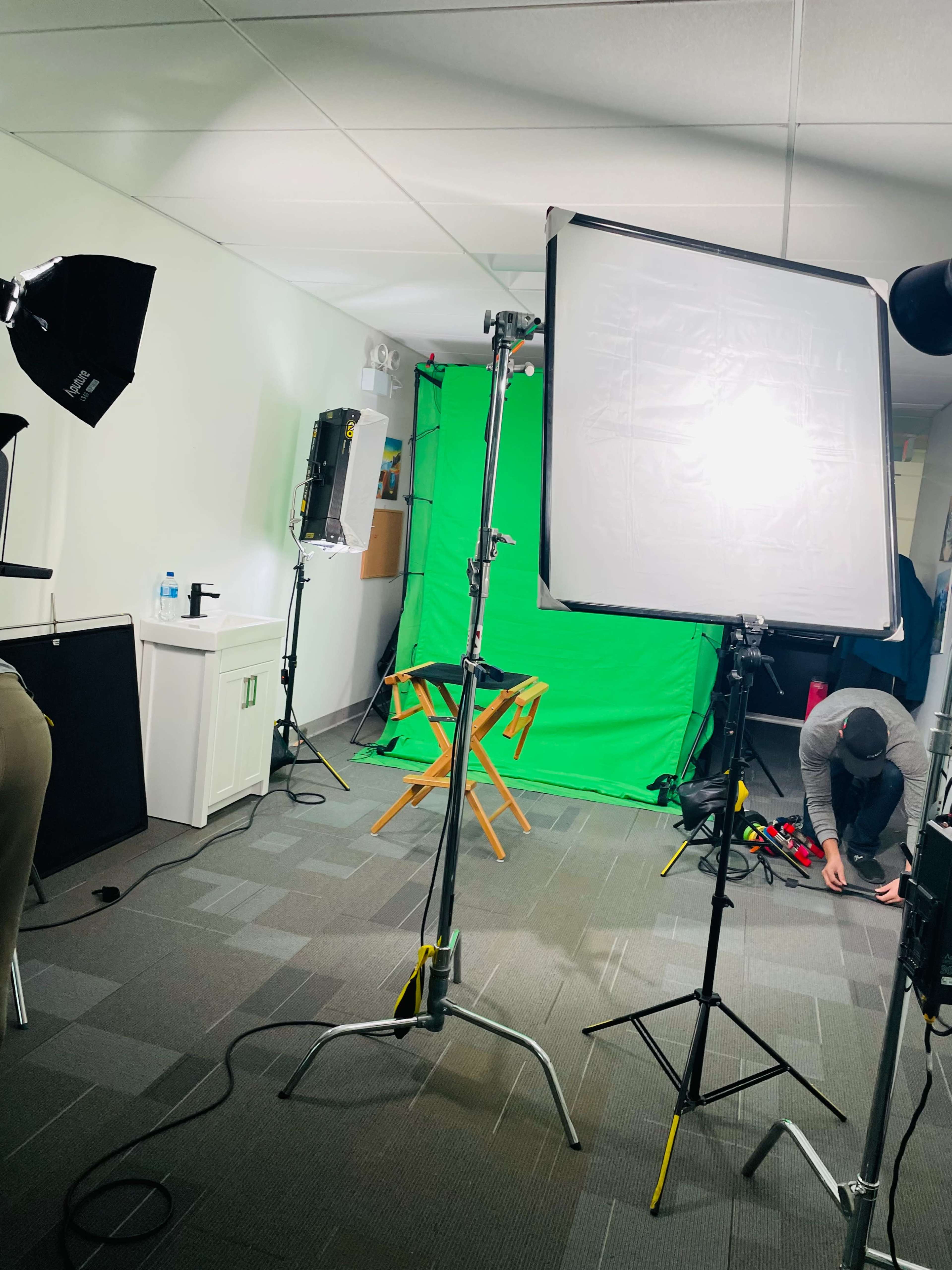A photo studio setup featuring a green screen backdrop, multiple light sources, and a director's chair, with a person adjusting equipment on the floor.