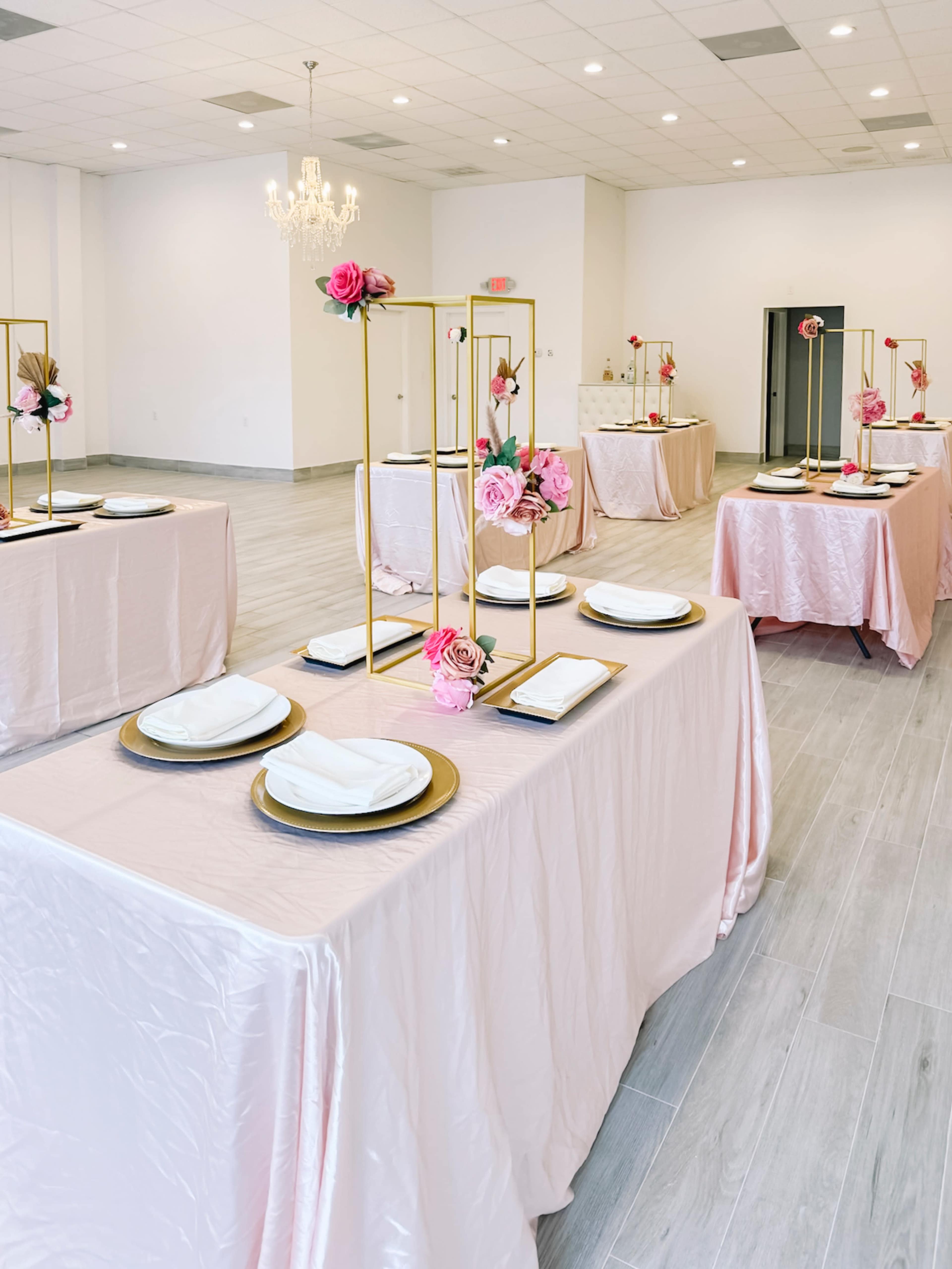 A banquet hall set up with tables covered in light pink cloths, adorned with elegant floral centerpieces and gold chargers.