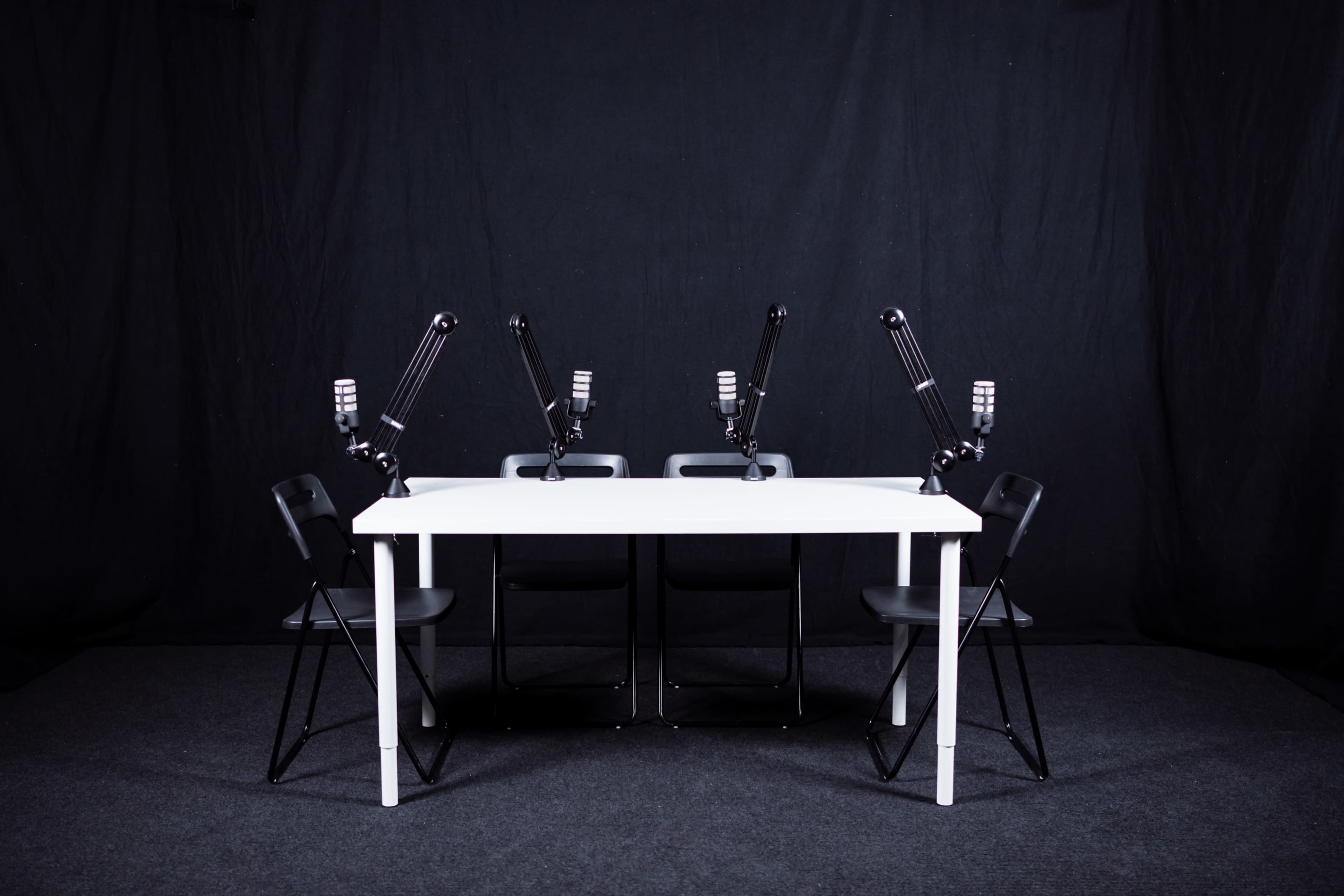 A white table with four black chairs and microphones mounted on adjustable arms is set against a black backdrop.