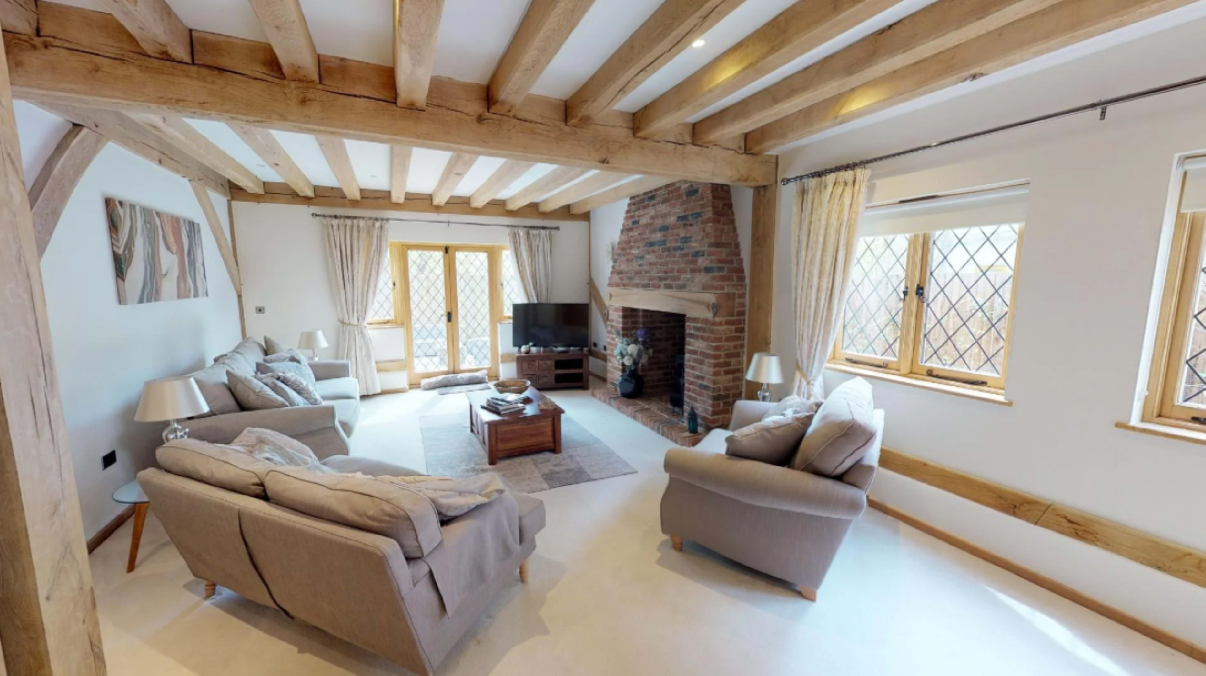 A spacious living room with exposed wooden beams, a brick fireplace, two sofas, a coffee table, and large windows letting in natural light.