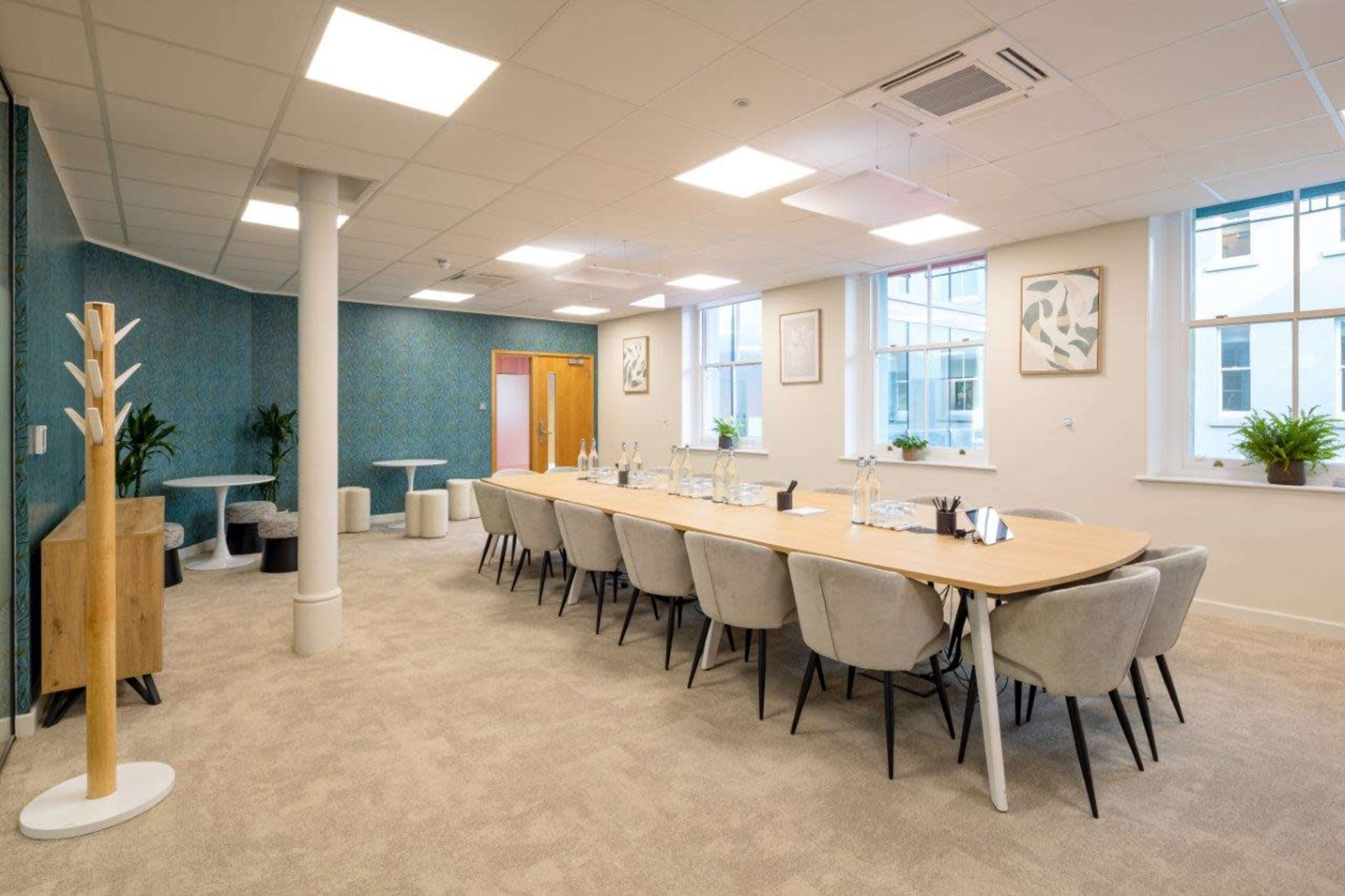 The image shows a modern conference room with a long wooden table surrounded by upholstered chairs, framed artwork on the walls, and two plants near the windows.