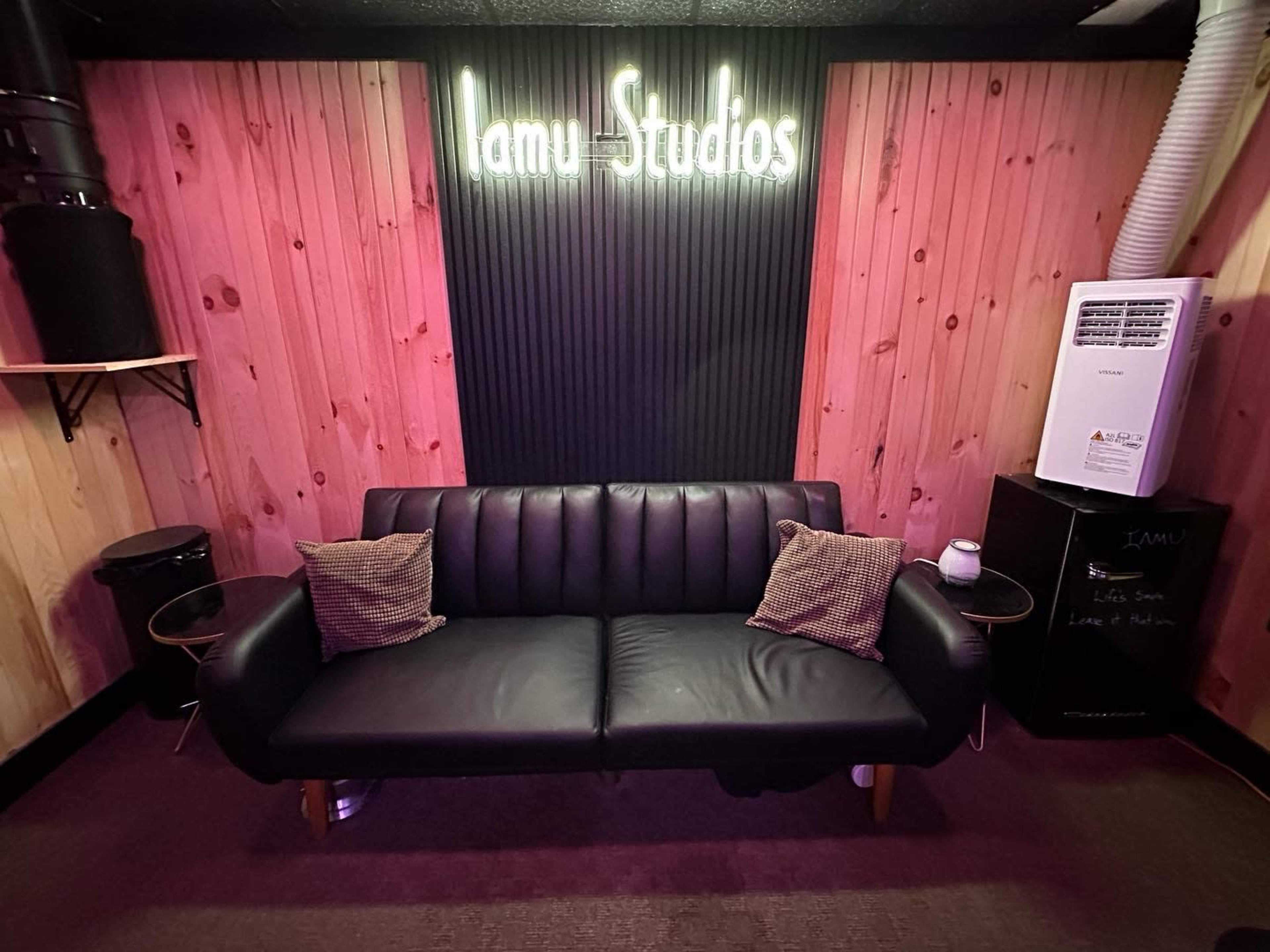 A black sofa with two pillows in front of a wooden wall featuring a neon sign that reads "Iamu Studios."