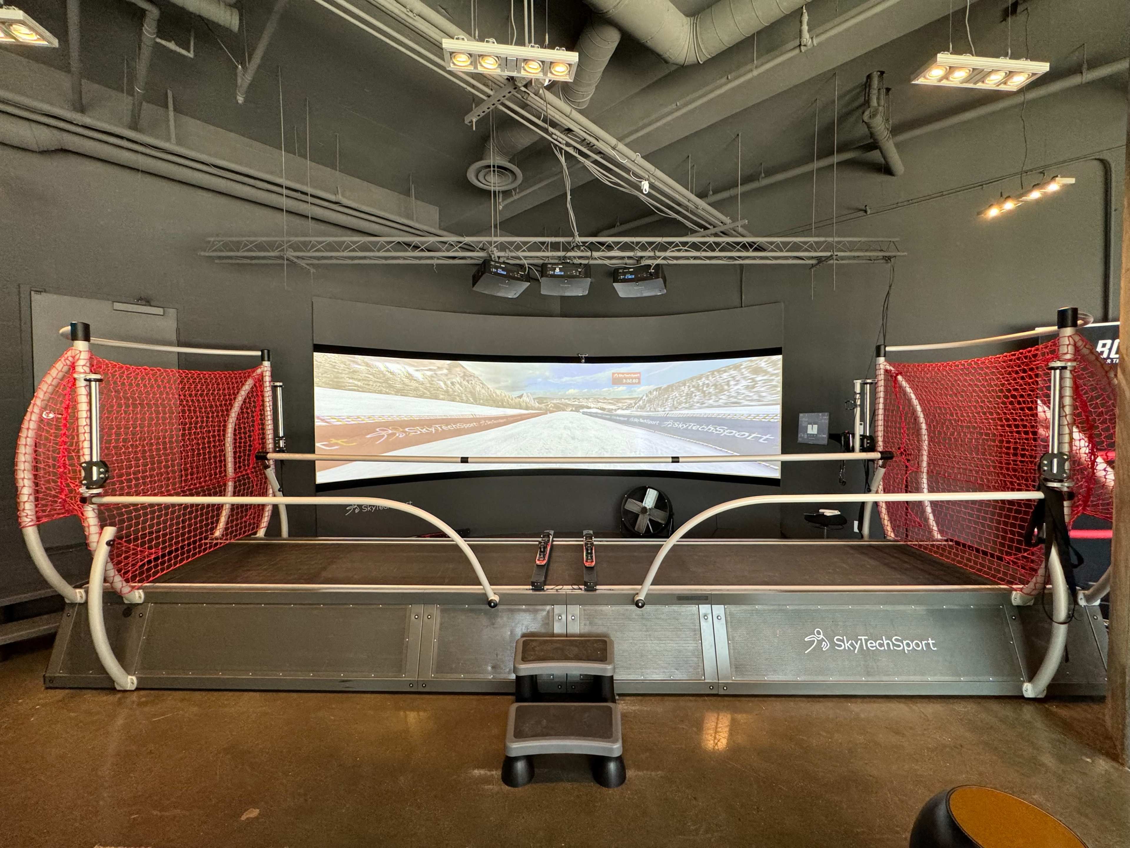 The image shows a sports simulation setup featuring a large screen displaying a winter landscape, with a central platform surrounded by red netting.