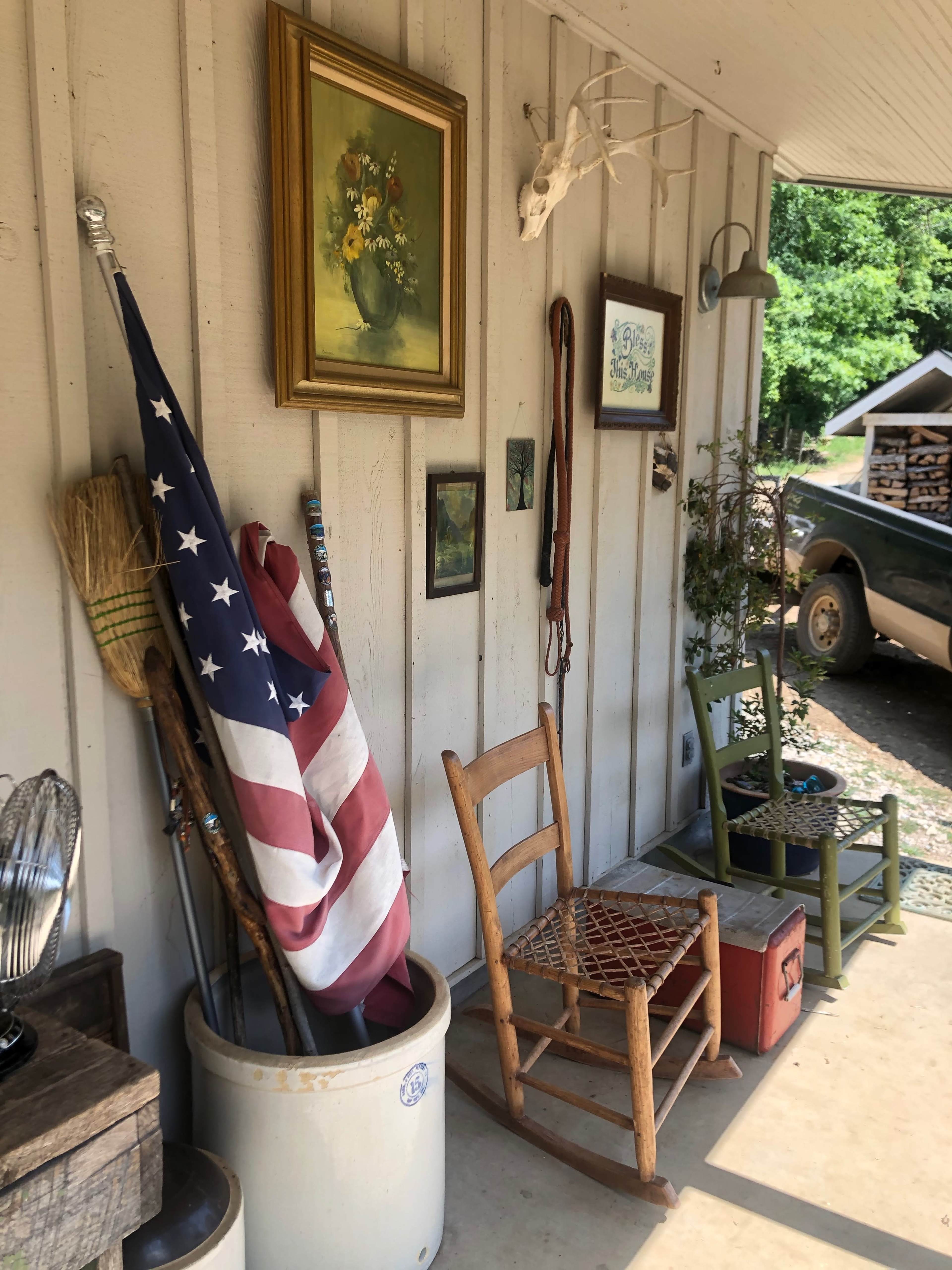 A rustic porch with an American flag, various decorations on the walls, a wooden rocking chair, and a green chair beside a vintage cooler.