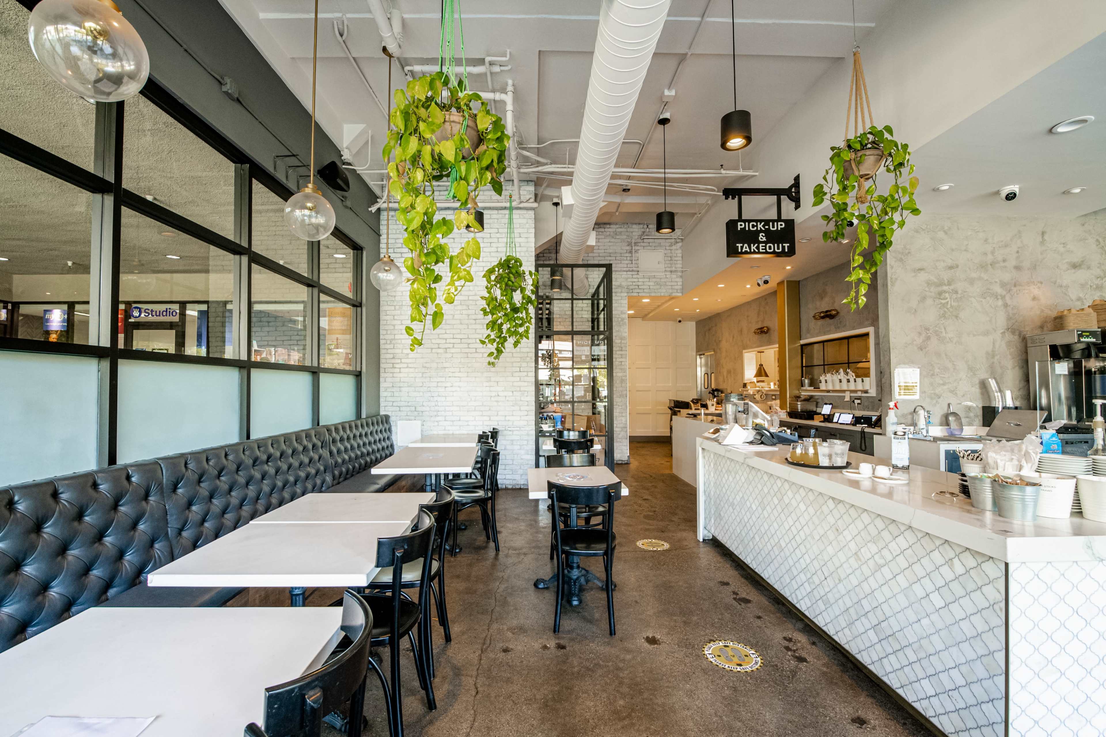 The image shows a modern cafe interior with black padded seating, white tiled counters, and hanging plants above the seating area.