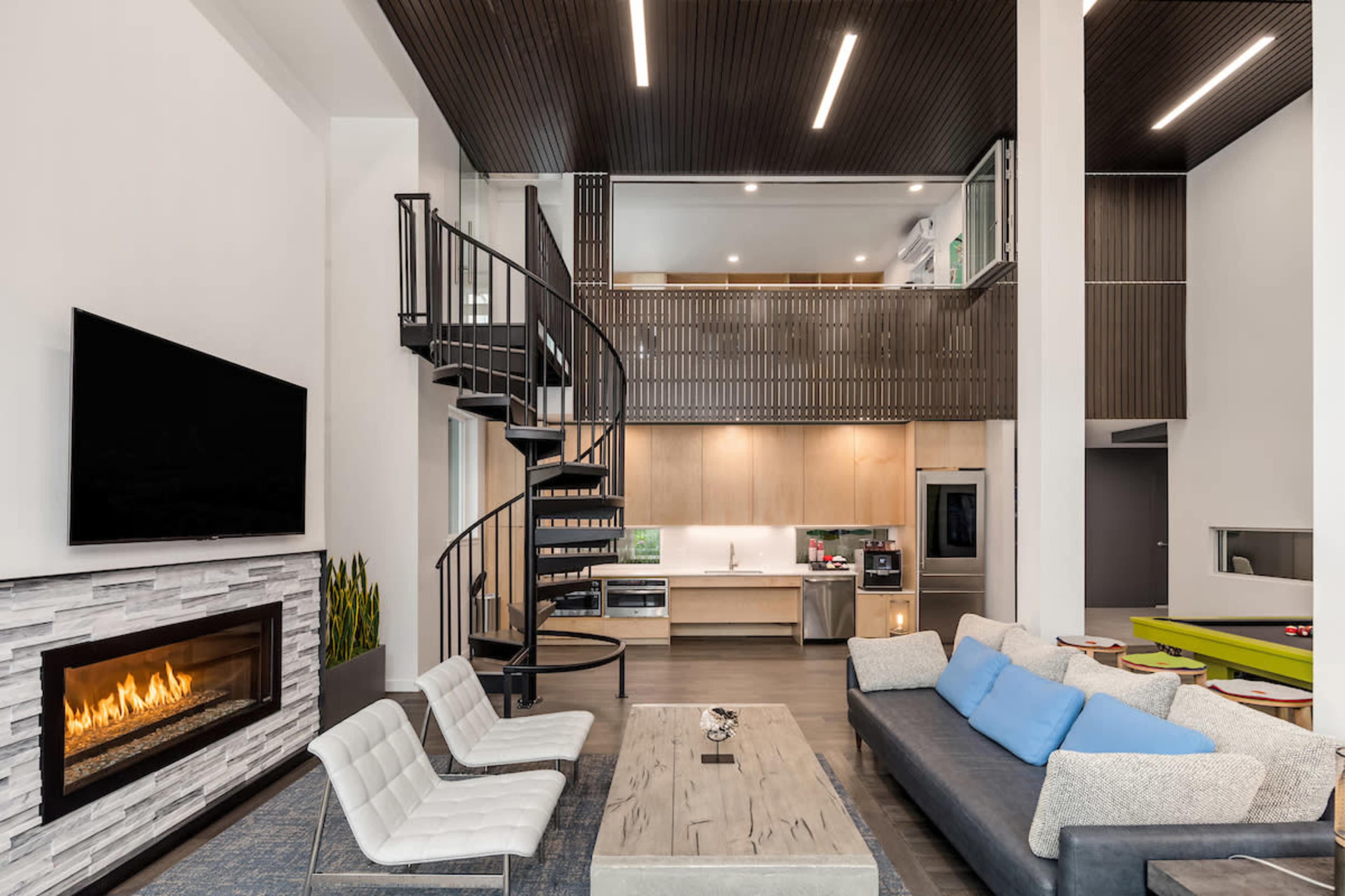 A modern living room with a spiral staircase, a large fireplace, and a kitchen visible in the background.