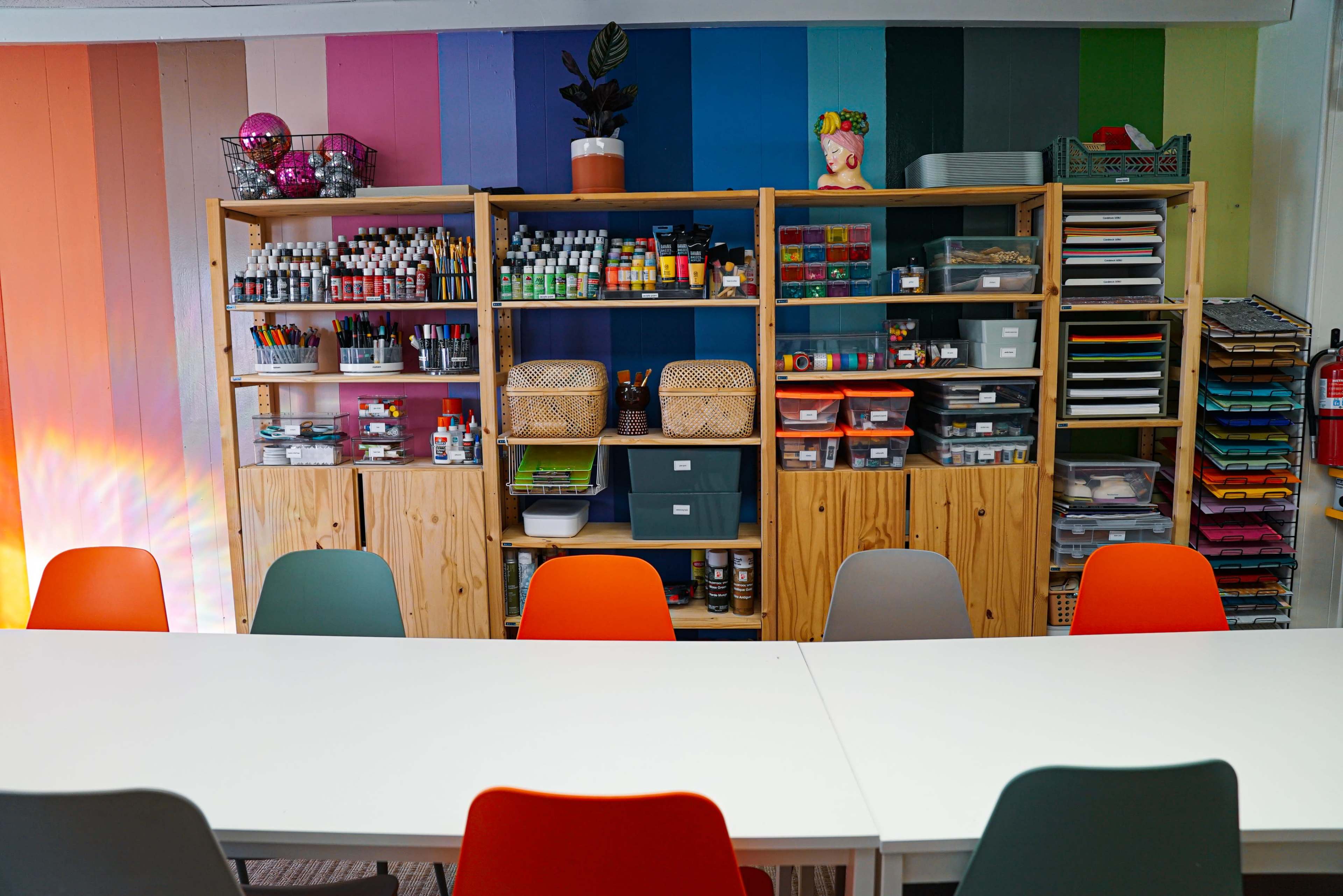 The image shows a brightly colored classroom or creative space with shelving units containing art supplies and a long white table surrounded by colorful chairs.