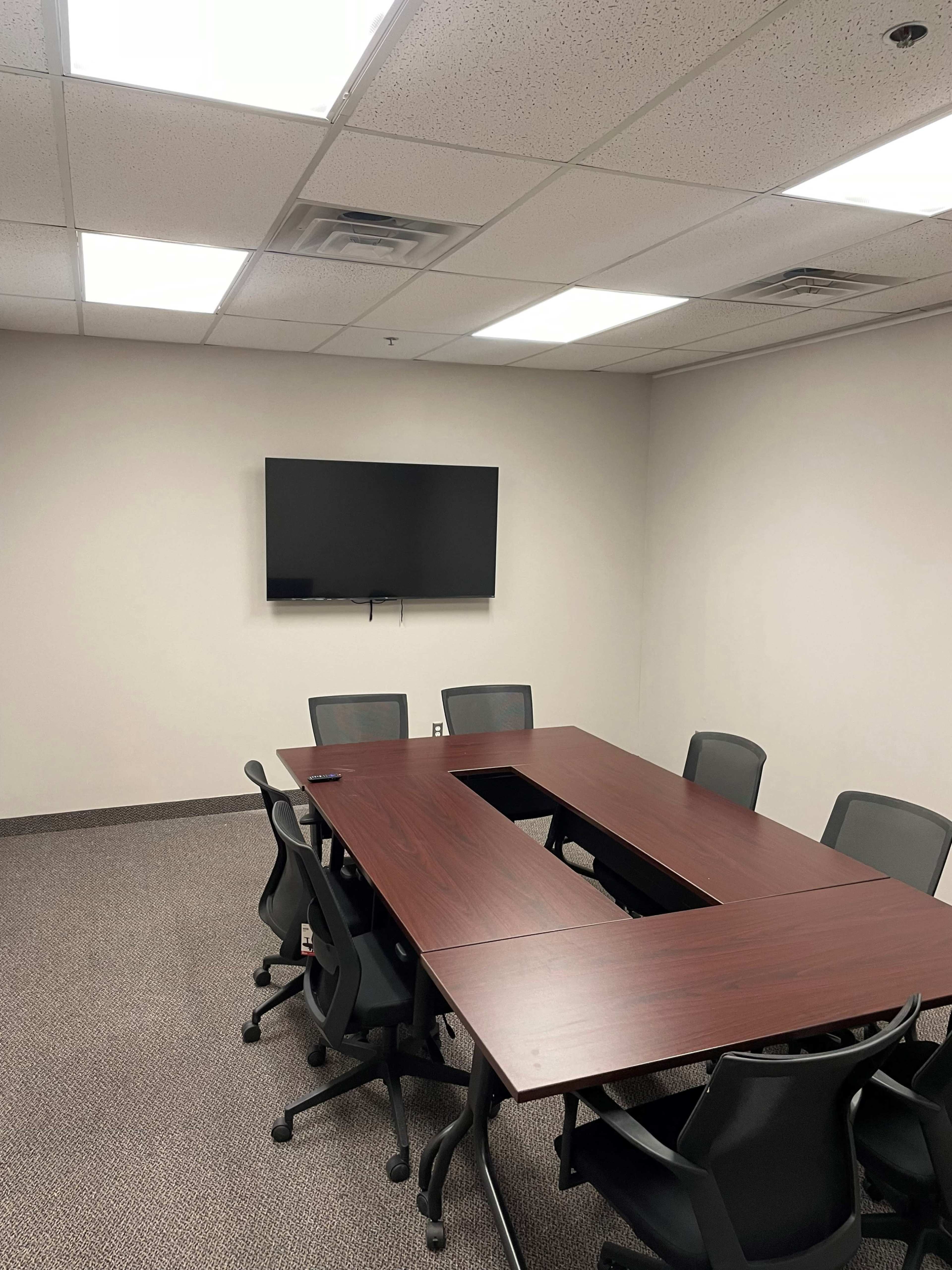 A conference room with a large wooden table surrounded by rolling chairs and a mounted television on the wall.