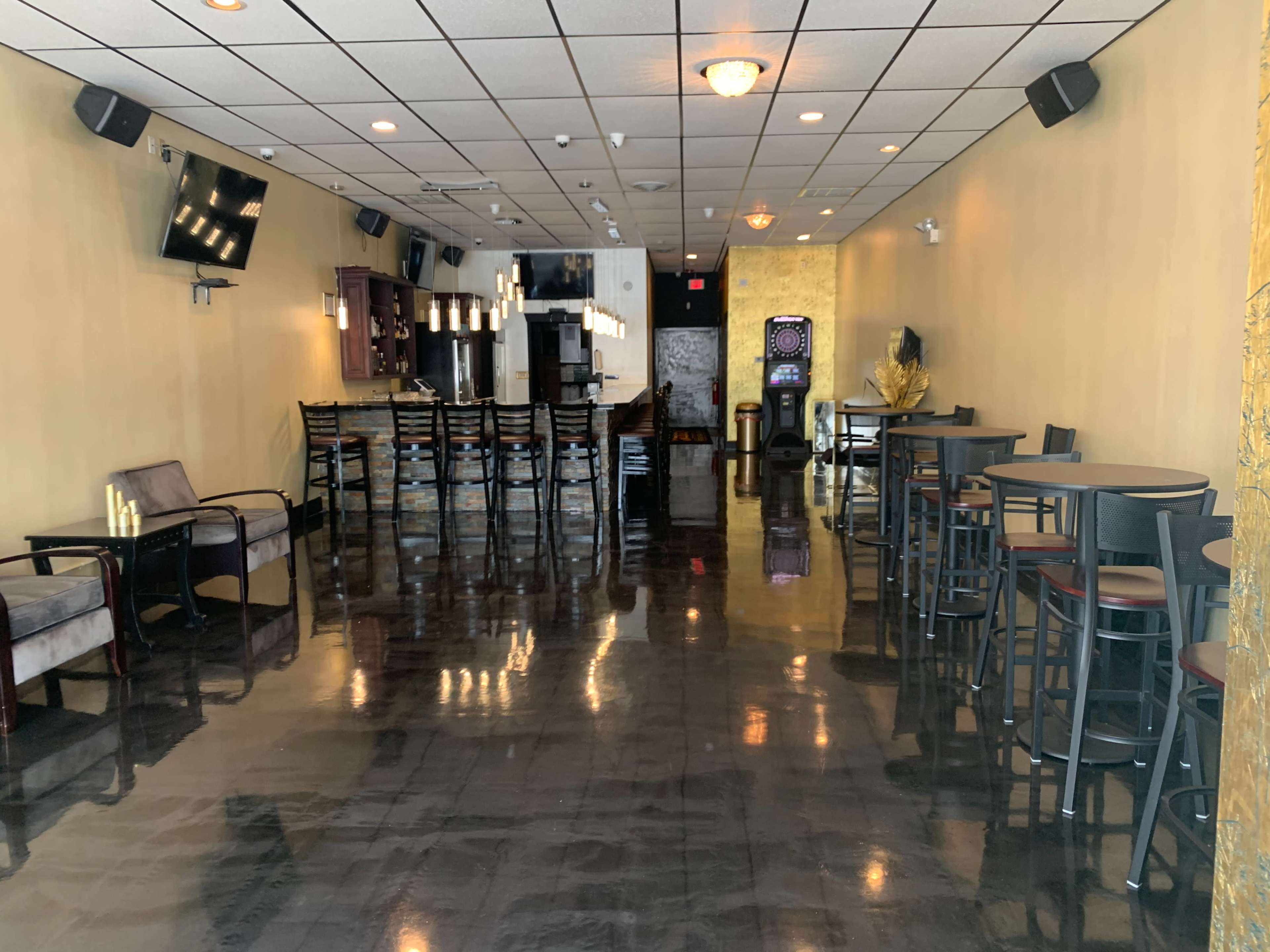 A spacious interior of a modern lounge or bar featuring black polished floors, overhead lighting, a long bar counter, and various seating arrangements.