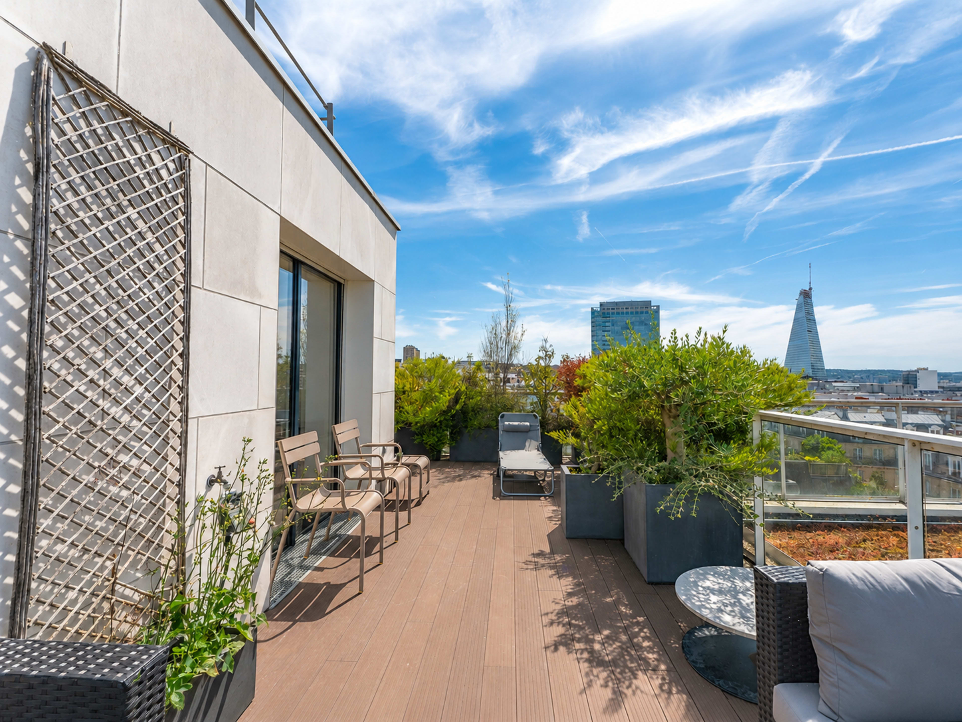 The image shows a modern rooftop terrace featuring seating, greenery, and a view of city buildings and a tower in the distance under a clear blue sky.