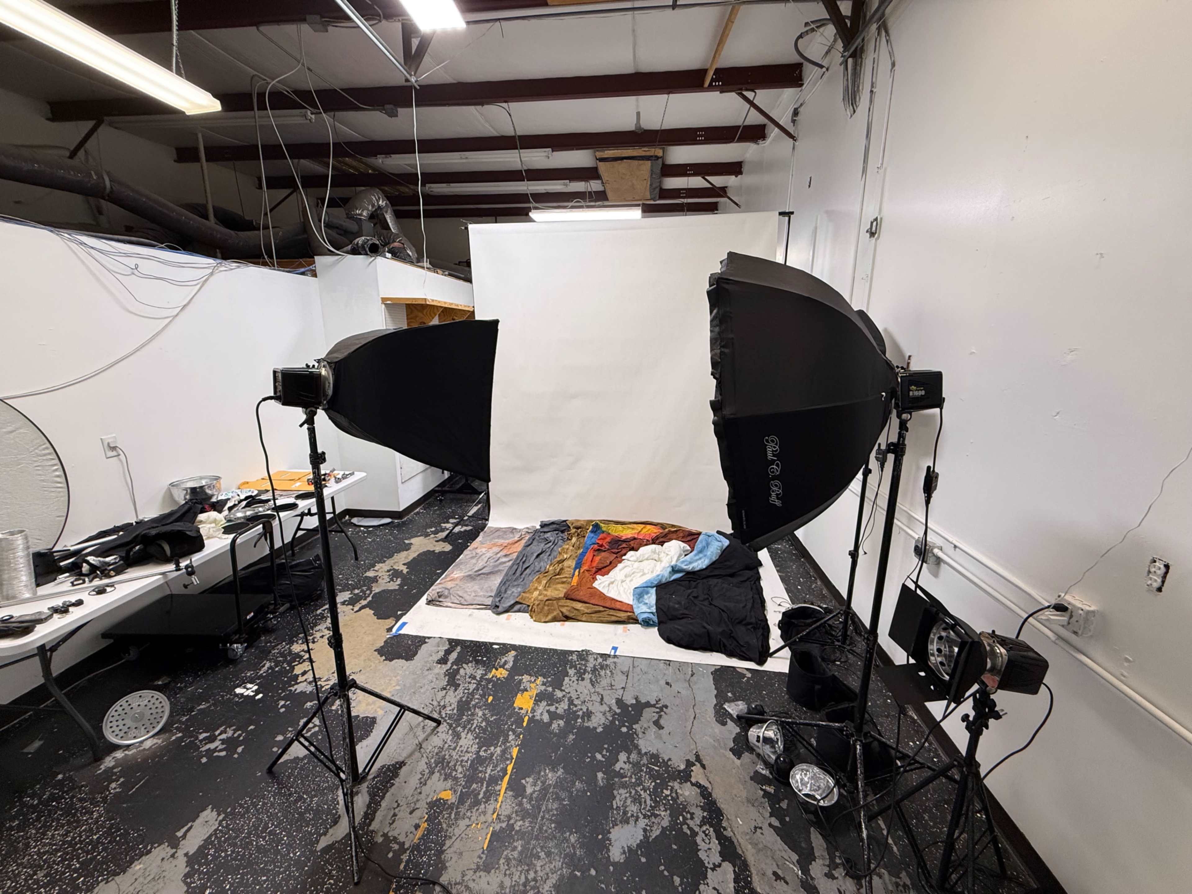 The image shows a photography studio setup with two softbox lights, a white backdrop, and a colorful assortment of fabrics laid out on the floor.