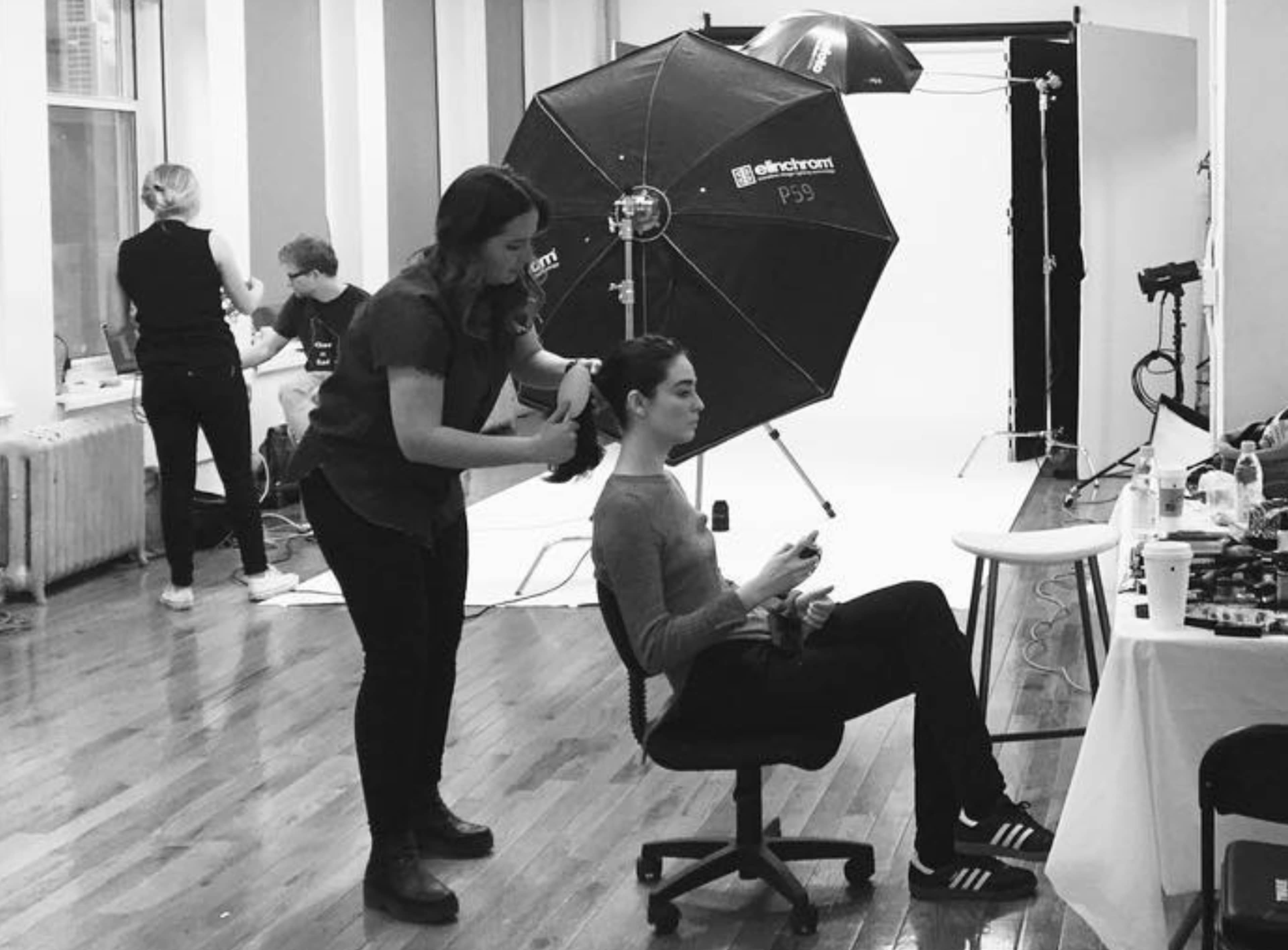 A person sits in a chair while another styles their hair in a photography studio with equipment set up in the background.