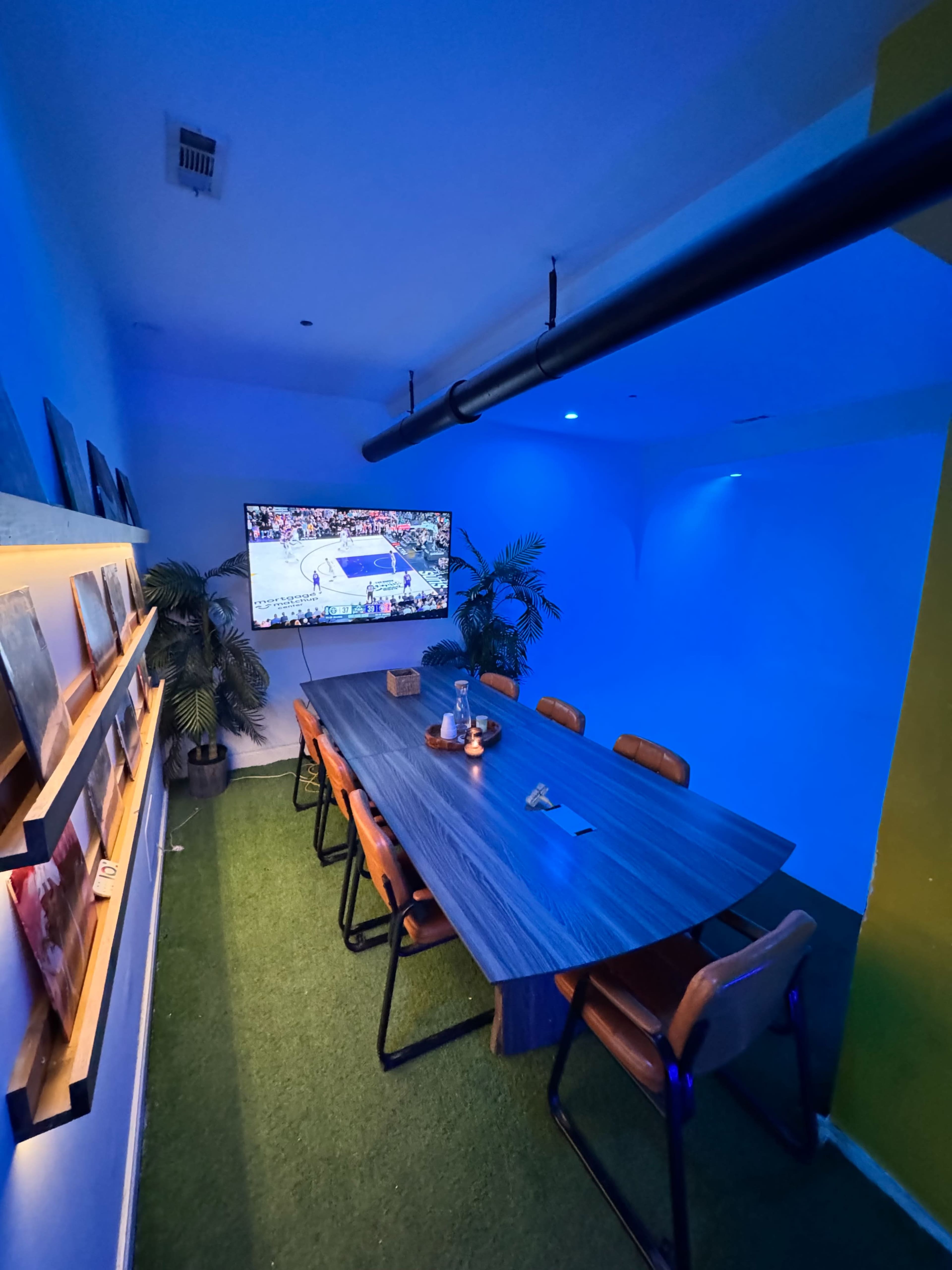 A conference room features a long table surrounded by chairs, a large screen showing a basketball game, and blue ambient lighting.