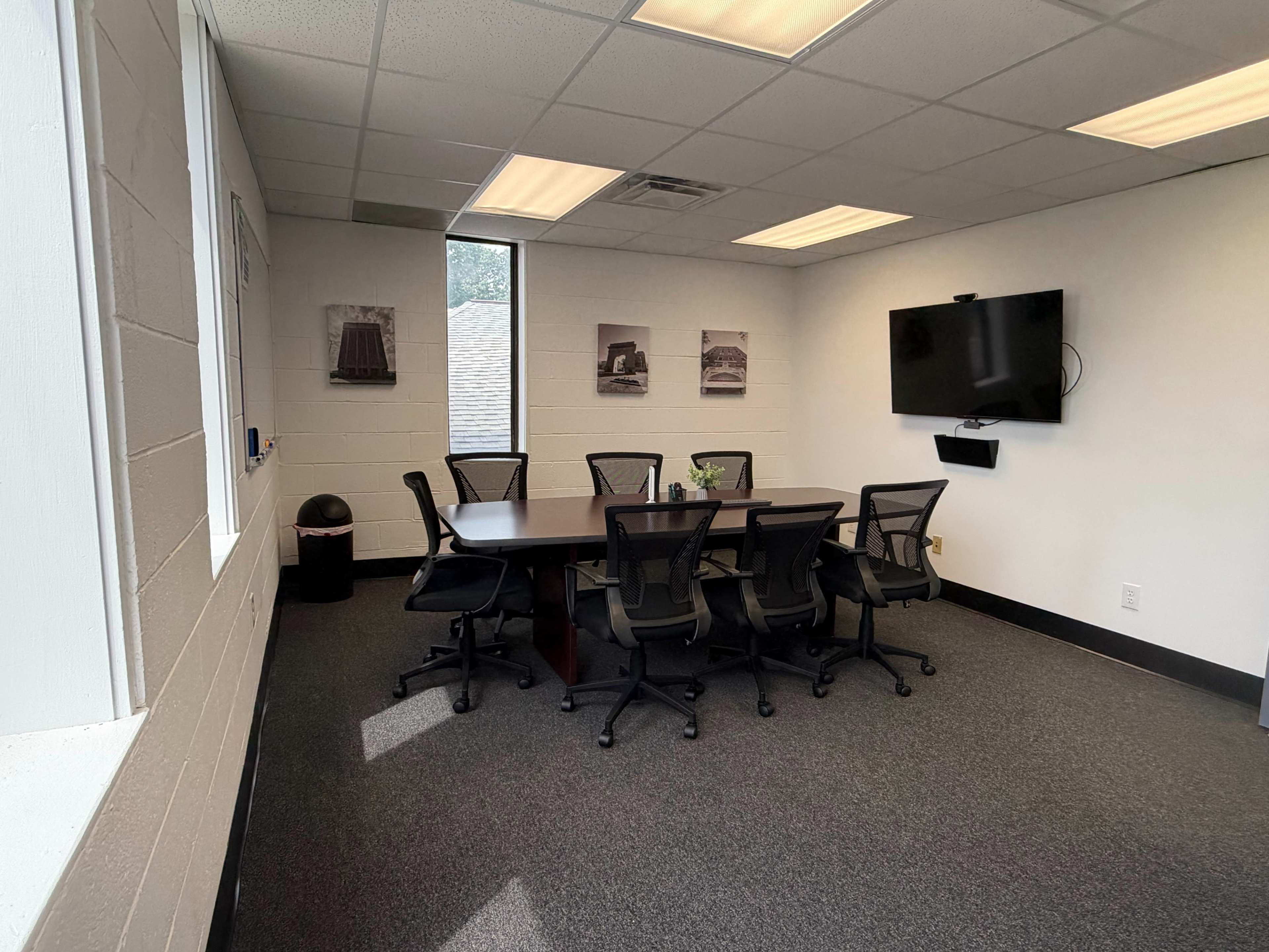 The image shows a conference room with a large table surrounded by several black office chairs, a television on the wall, and framed photographs displayed on the adjacent walls.