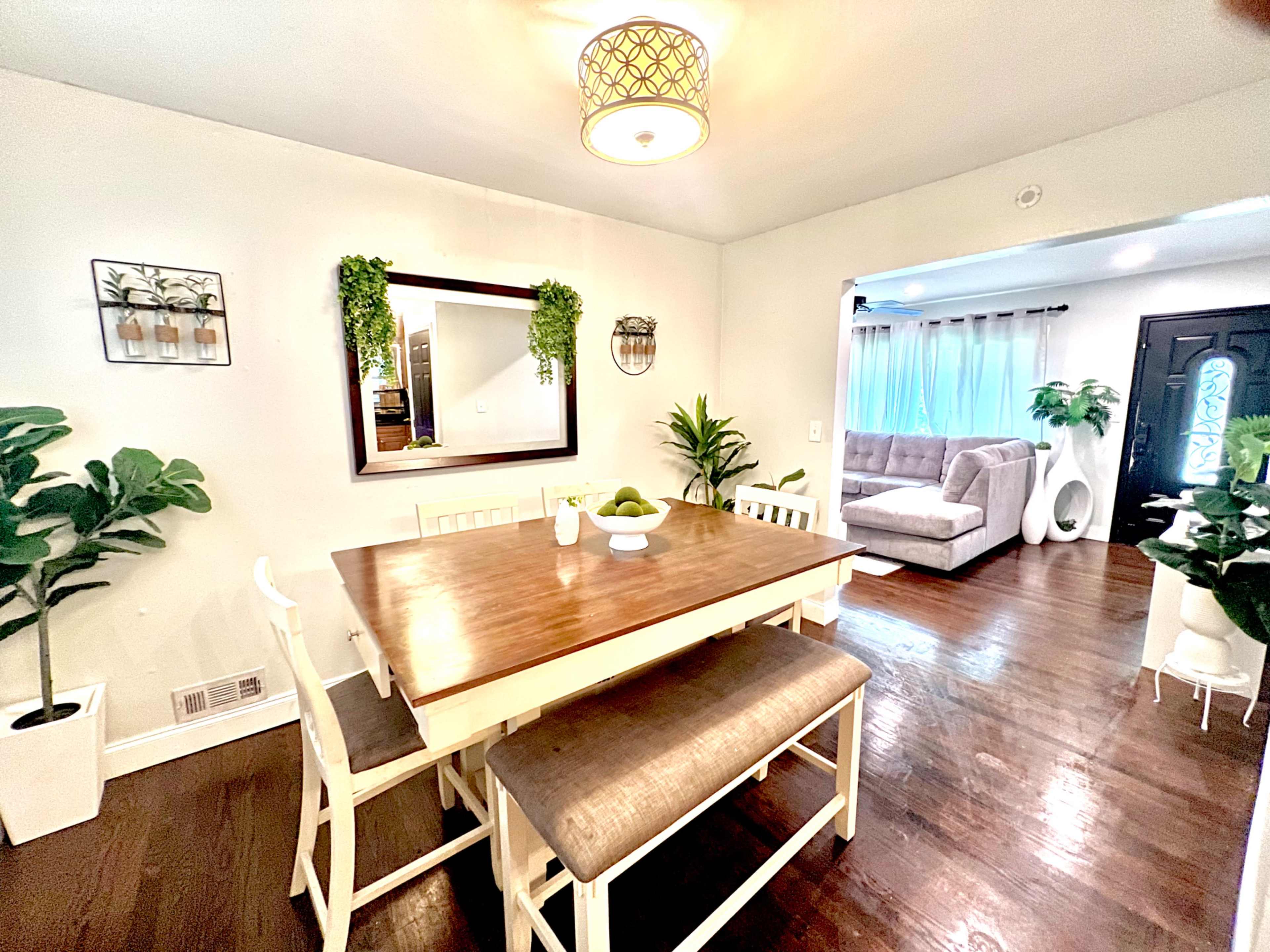 A dining area features a wooden table with white chairs, surrounded by potted plants and a large mirror.
