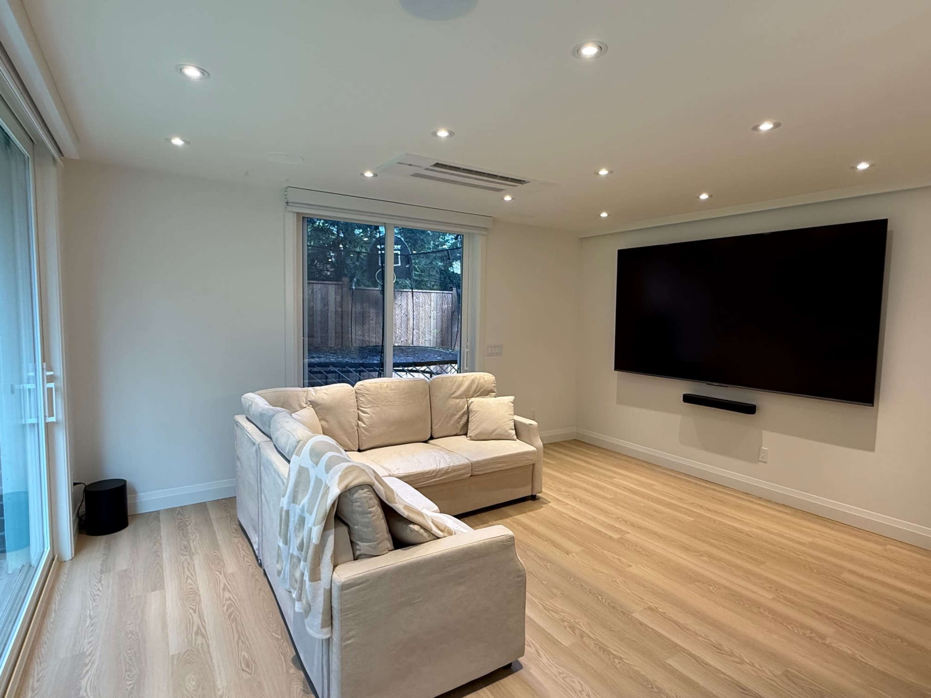 The image shows a modern living room featuring a white sofa, a large wall-mounted television, and a window overlooking a wooden fence.