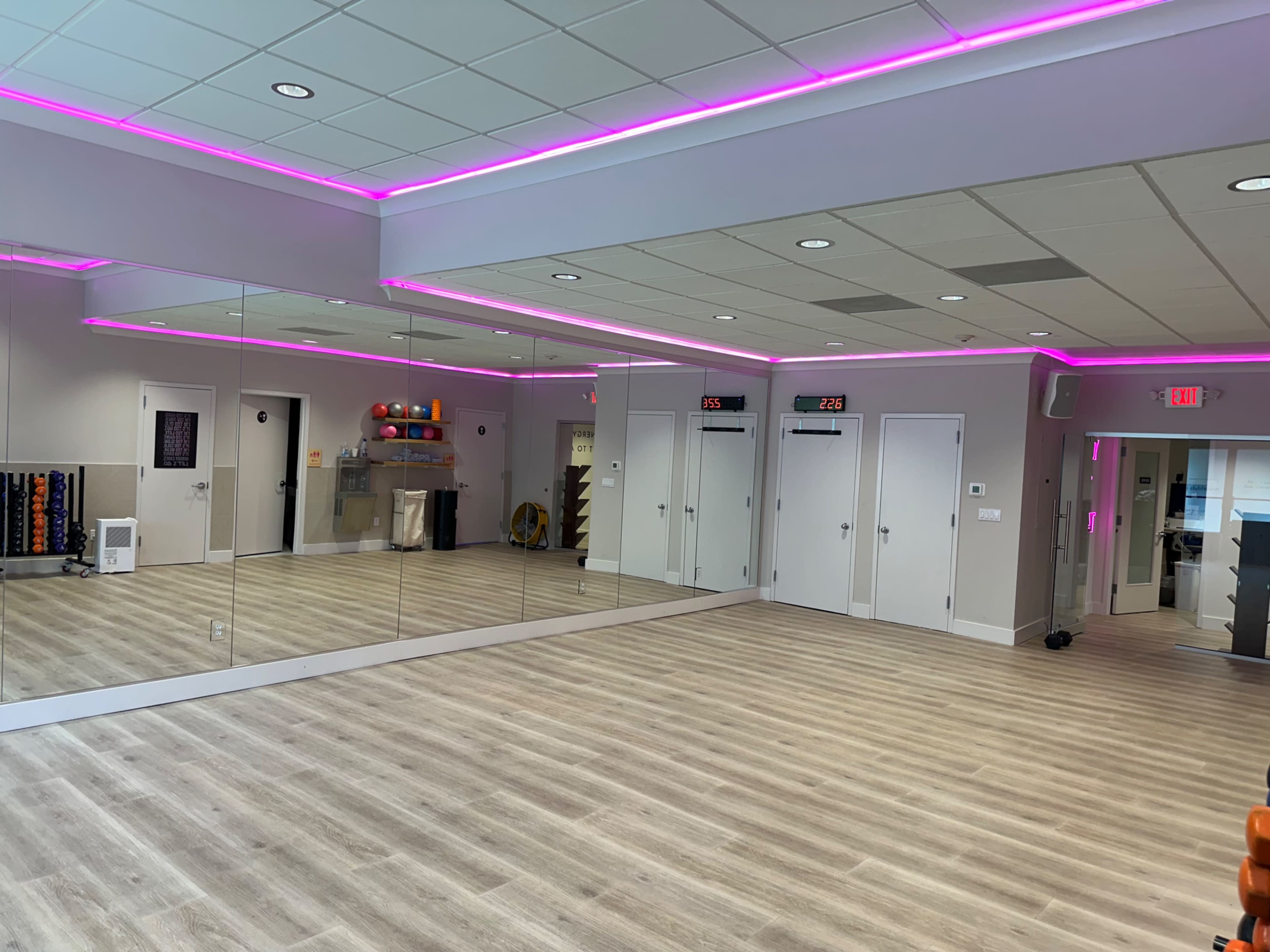 The image shows a well-lit fitness studio with mirrored walls, wooden flooring, and pink LED lighting along the ceiling.