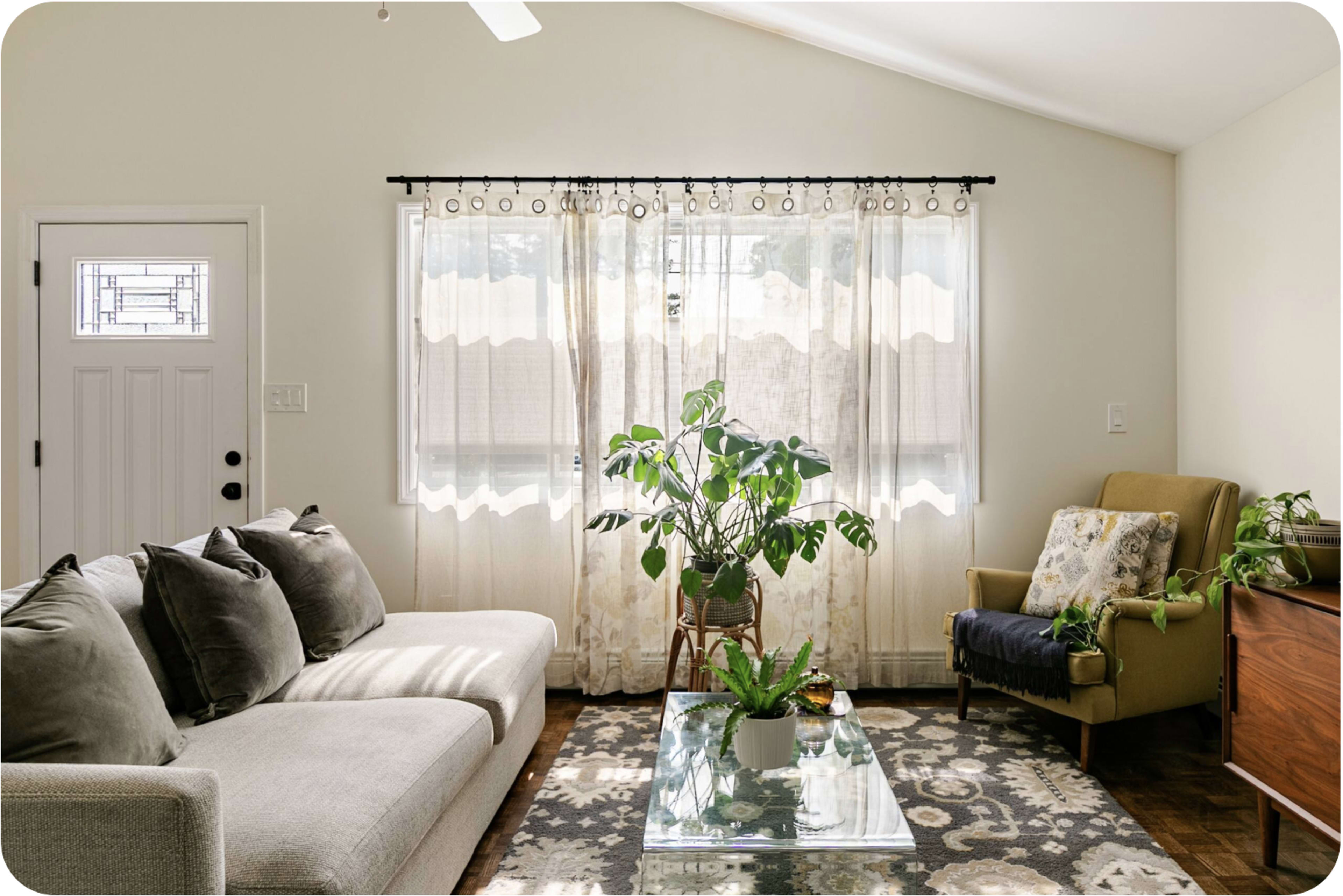 A light-colored living room features a sofa, an armchair, a glass coffee table, and plants, with sheer curtains allowing natural light to enter.