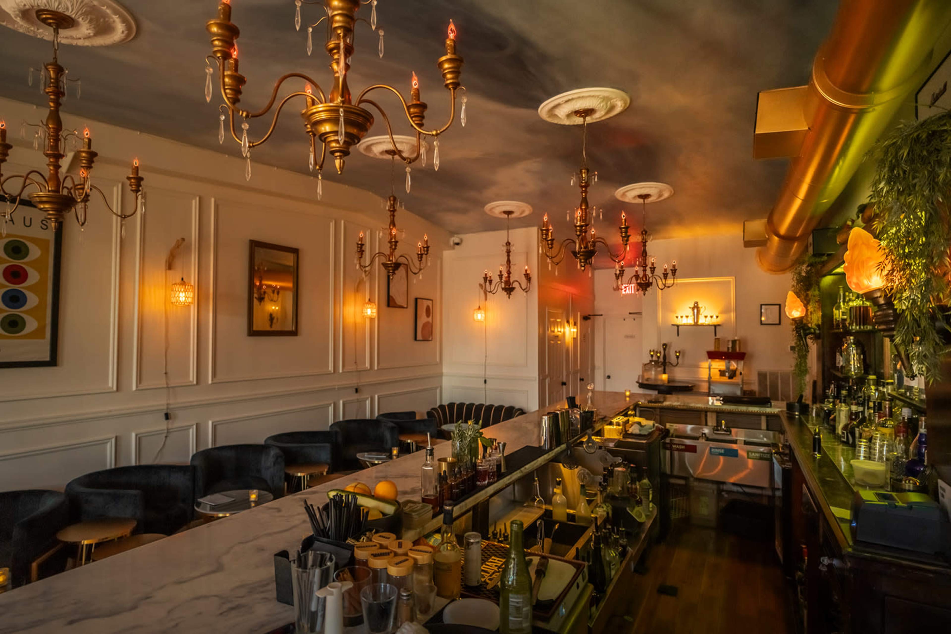 The interior of a dimly lit bar features a marble countertop, elegant chandeliers, and a well-stocked back shelf with various bottles.