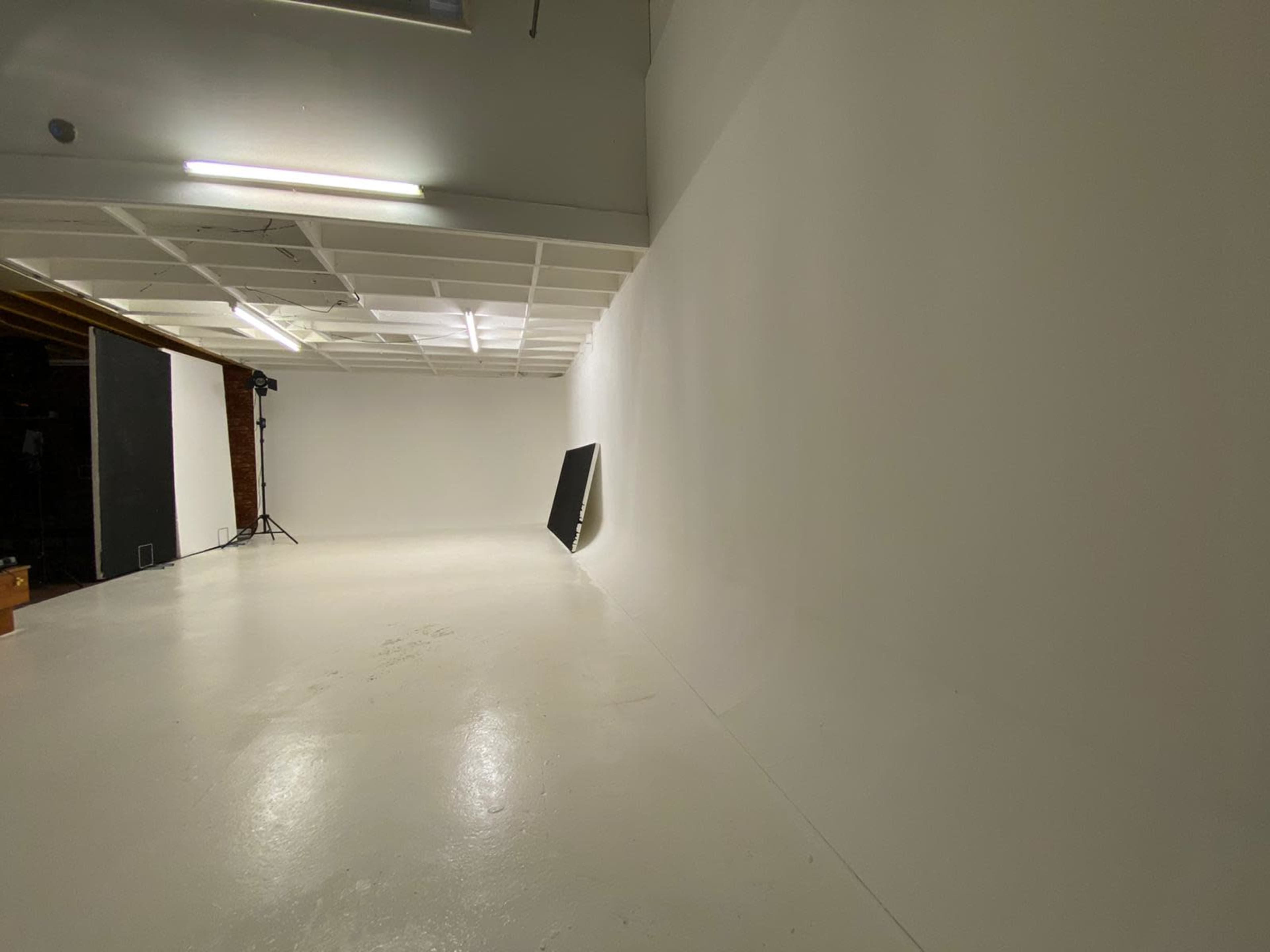 A spacious, empty studio with white walls and a polished floor, featuring a backdrop stand and soft lighting equipment in the corner.