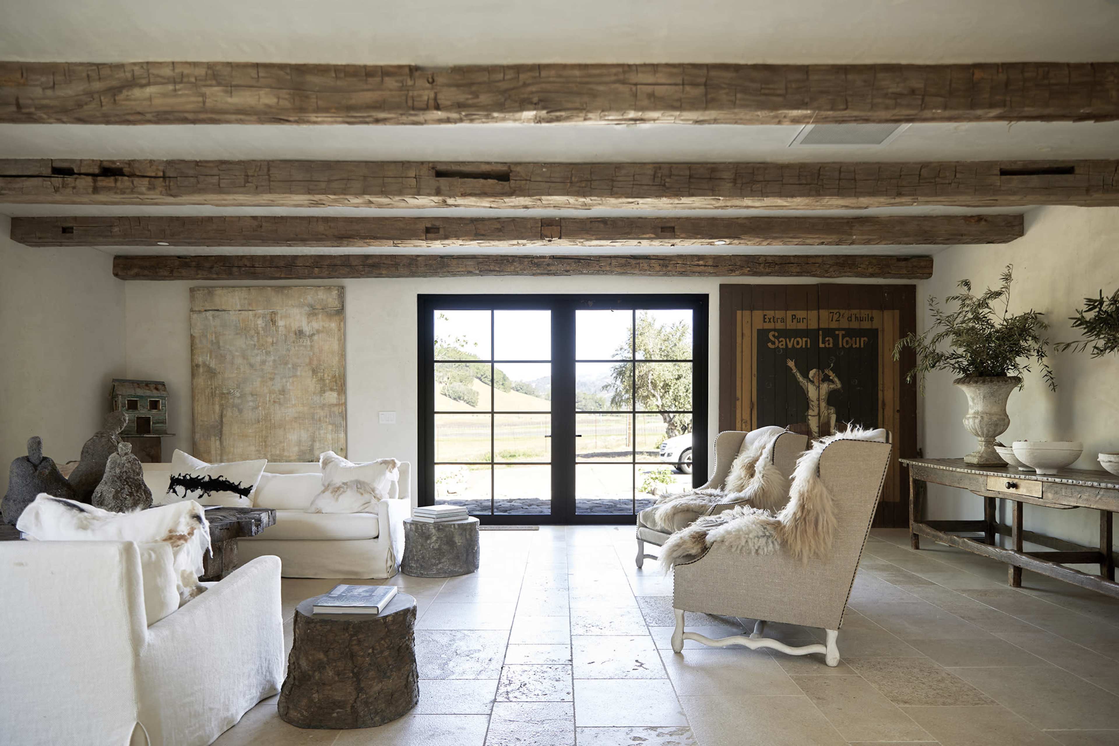 A spacious living room features wooden beams, stone flooring, and large windows overlooking a scenic landscape.