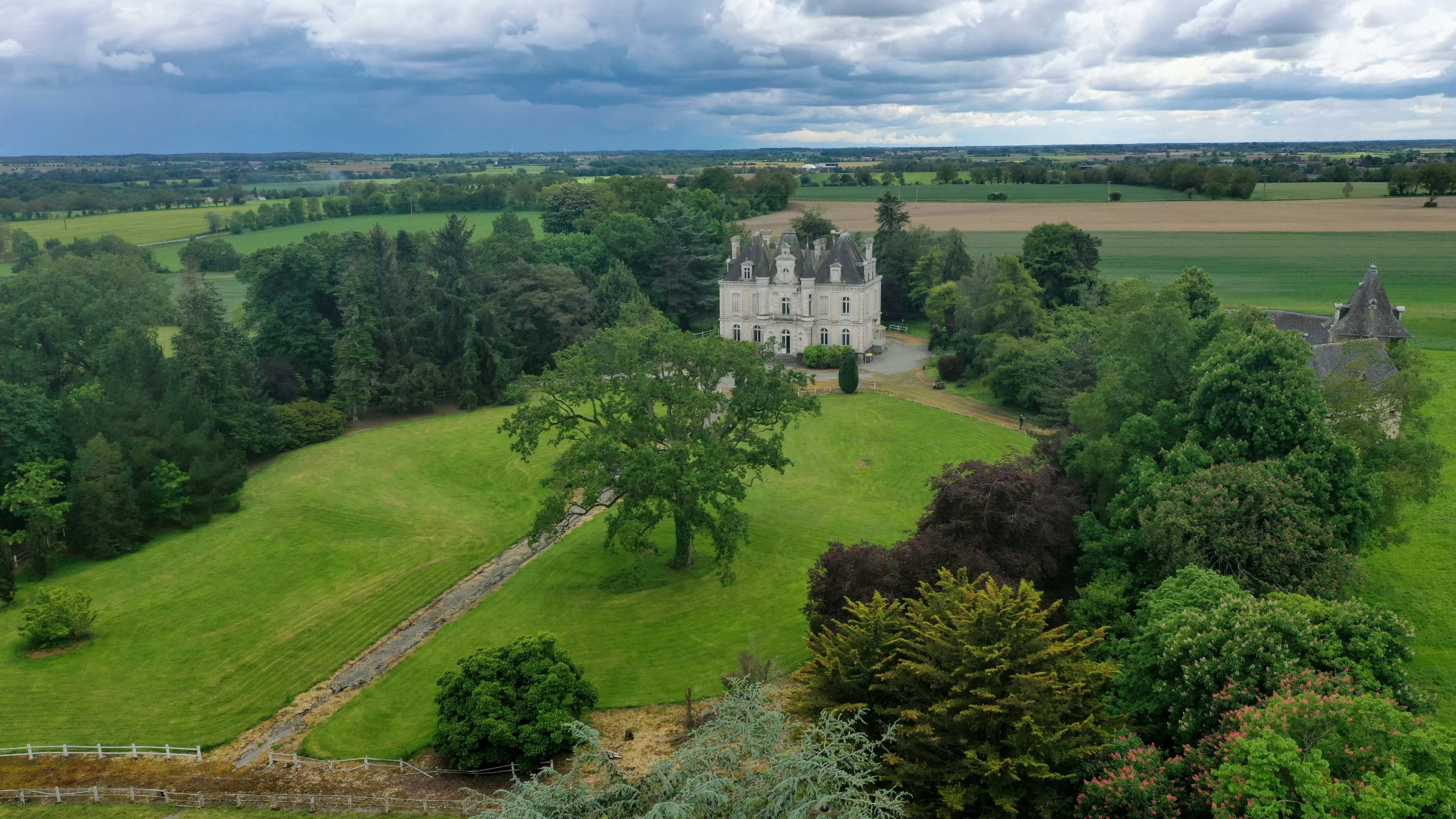 A grand, two-story mansion surrounded by lush greenery sits amidst a vast expanse of rolling fields under an overcast sky.