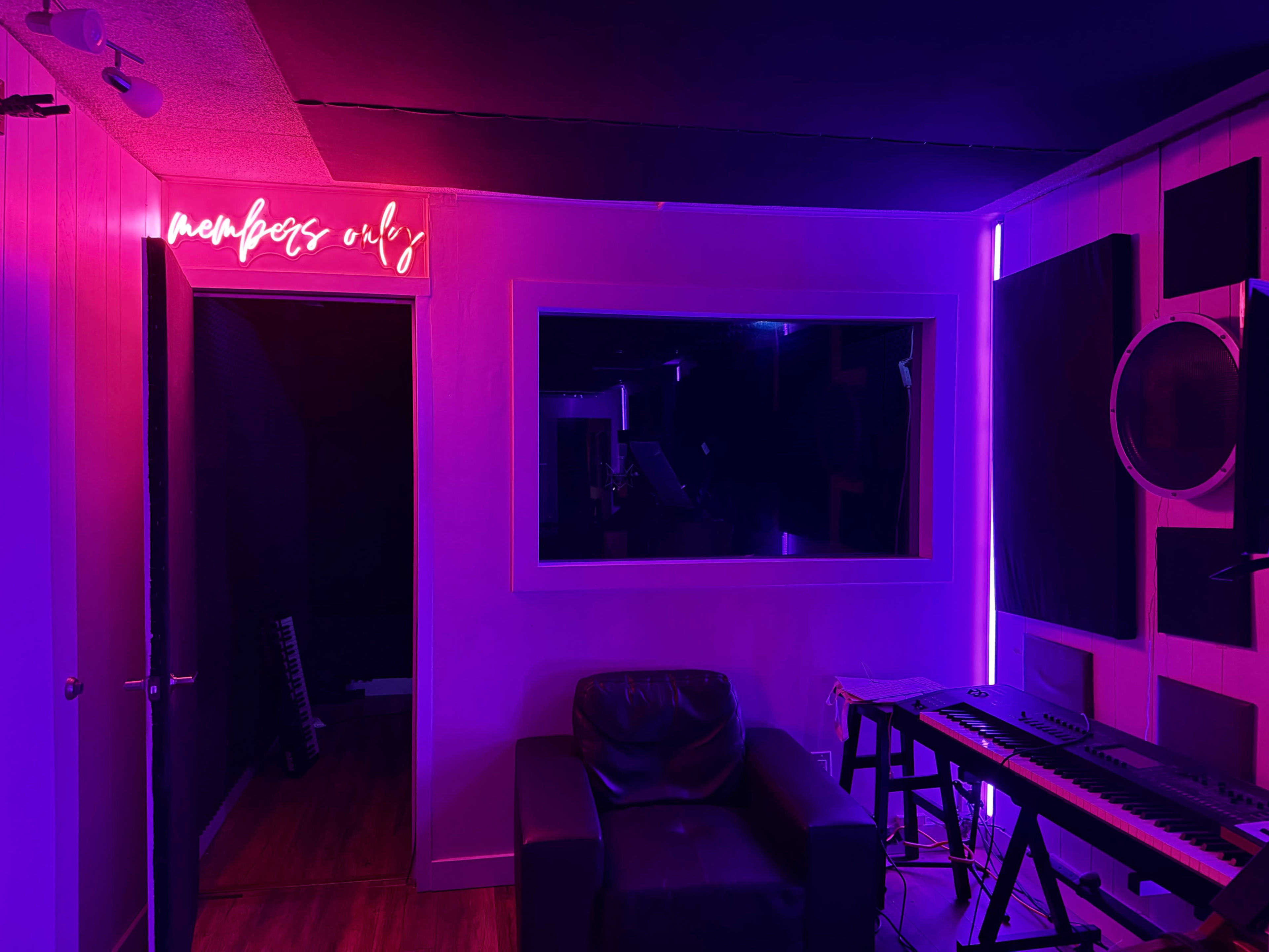 The image shows a music studio with neon lighting, a black leather chair, and a keyboard positioned near a mirror and an entrance labeled "members only."