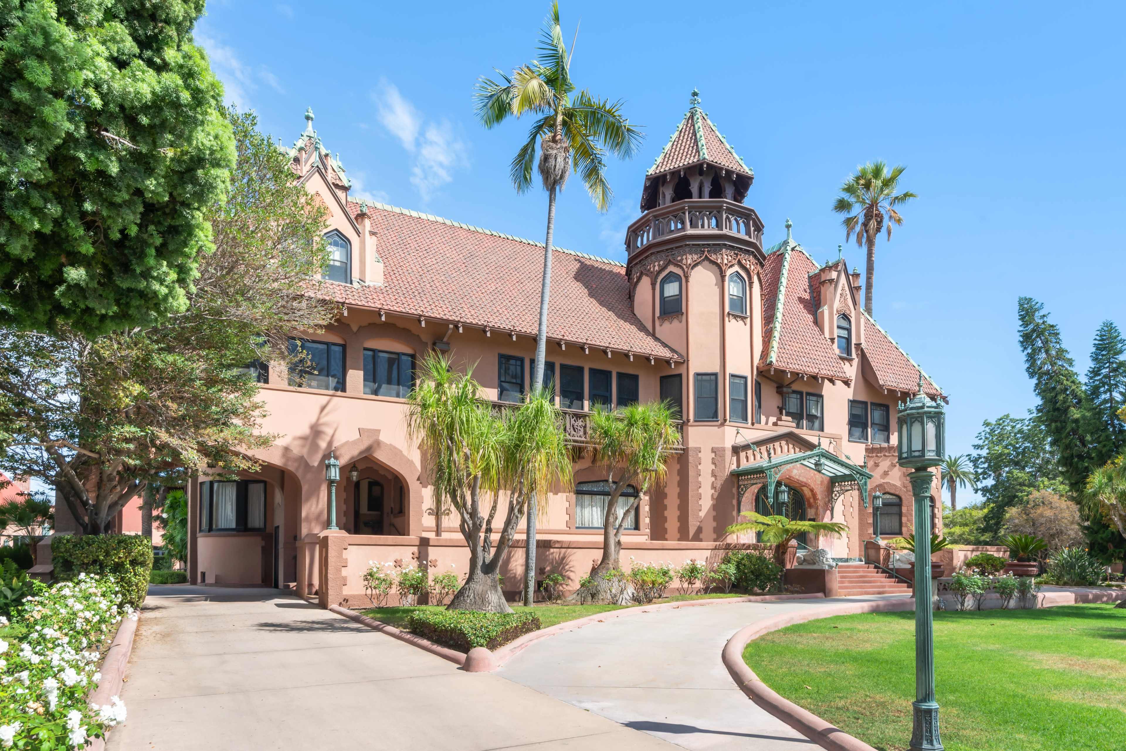 The image shows a large, ornate Victorian-style building with a turret, palm trees, and a pathway leading to the entrance.
