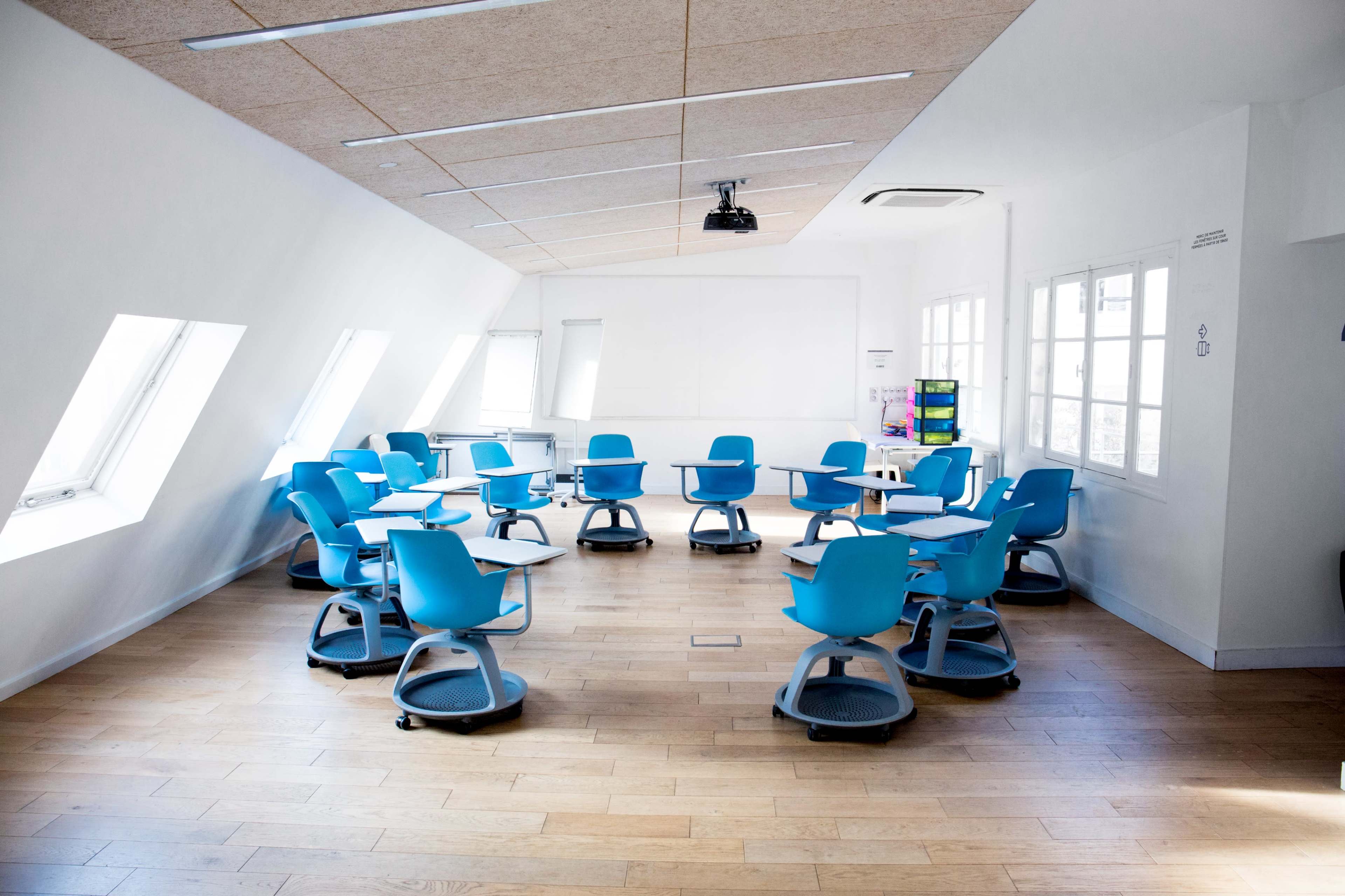A modern classroom features circular tables and blue rolling chairs arranged around the room with large windows and a ceiling-mounted projector.