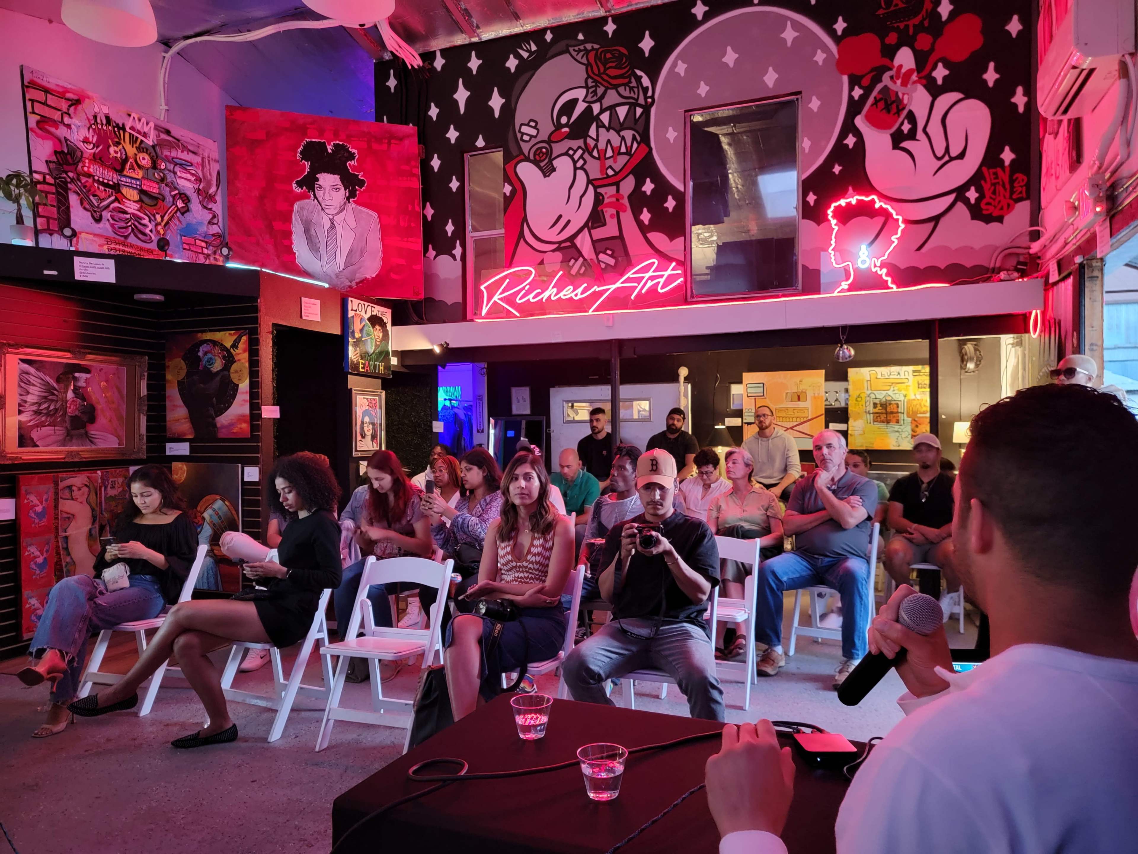 A crowd of people sits in wooden chairs inside an art gallery featuring colorful murals and neon signs, while a speaker addresses them from the front.