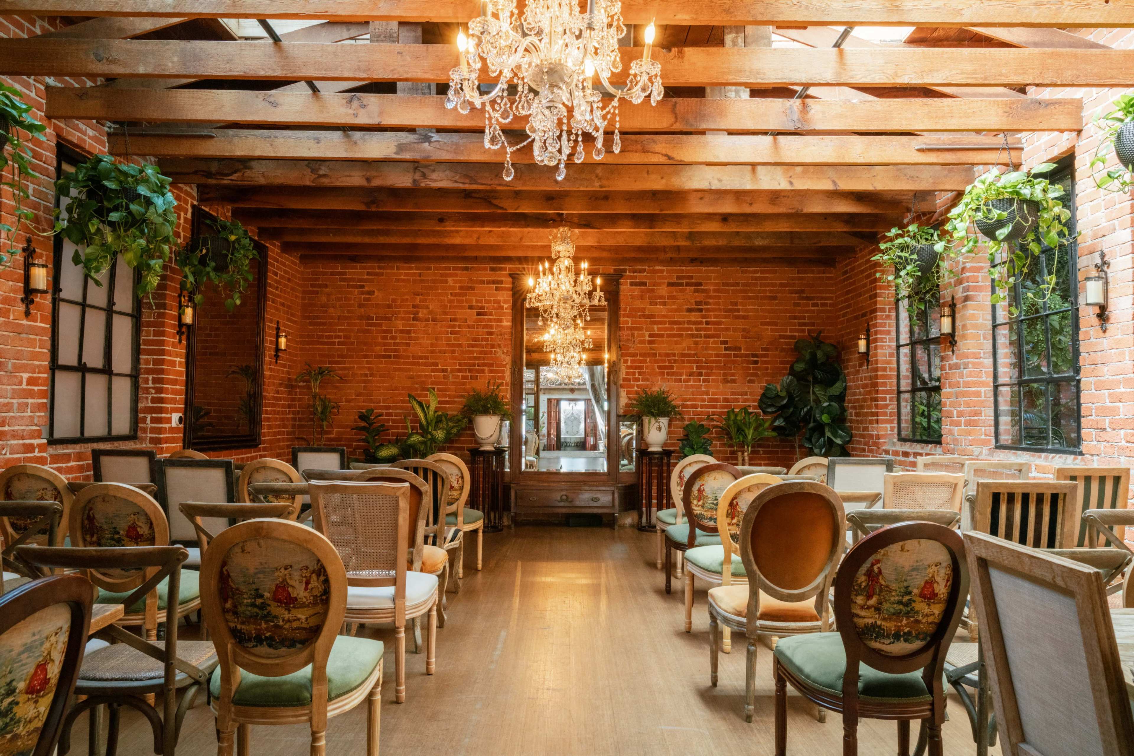 The image shows a well-lit room with wooden beams, exposed brick walls, and rows of vintage chairs accented by chandeliers and potted plants.