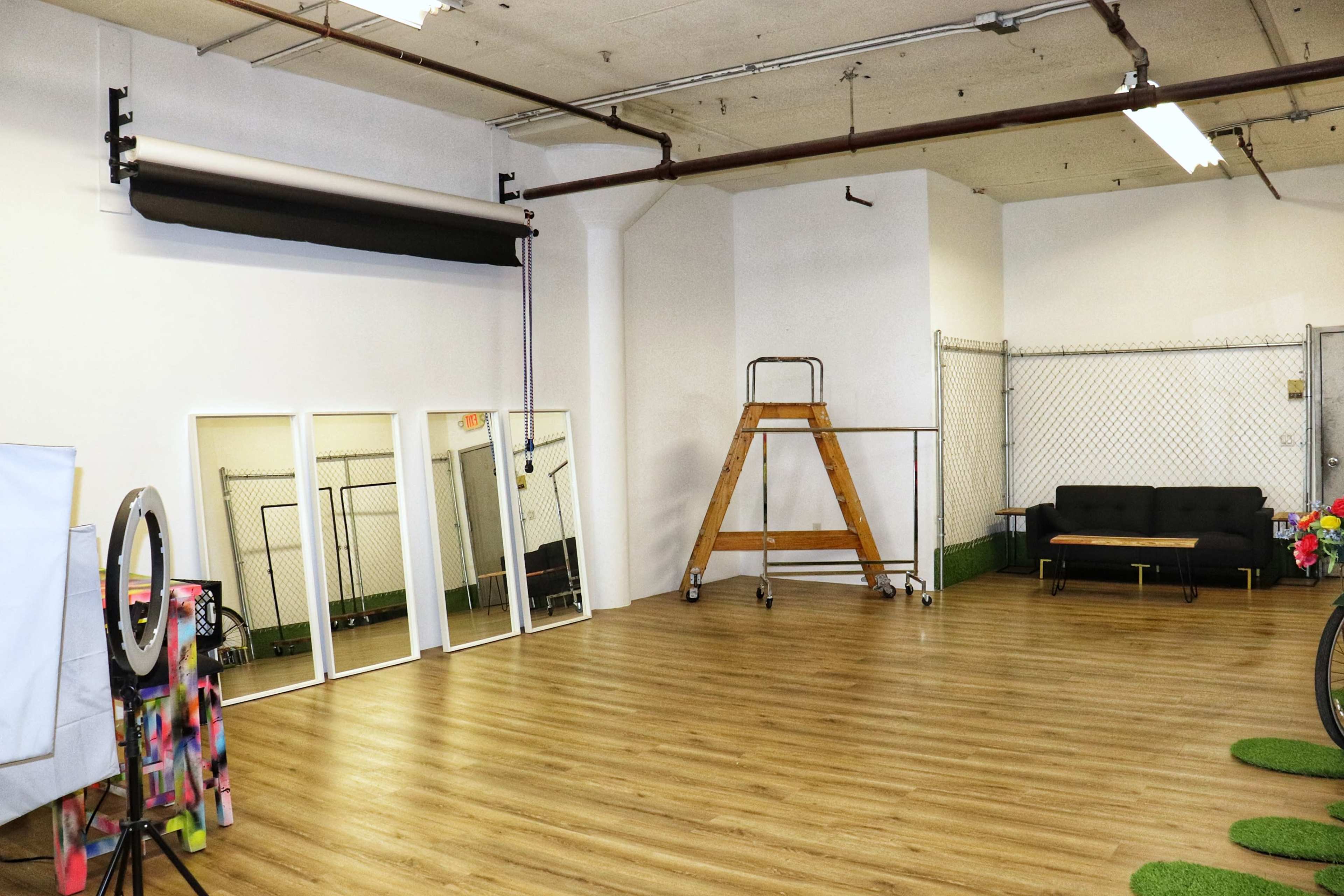 A spacious room with wooden flooring, featuring a black couch, a folding ladder, mirrors, and a backdrop setup.