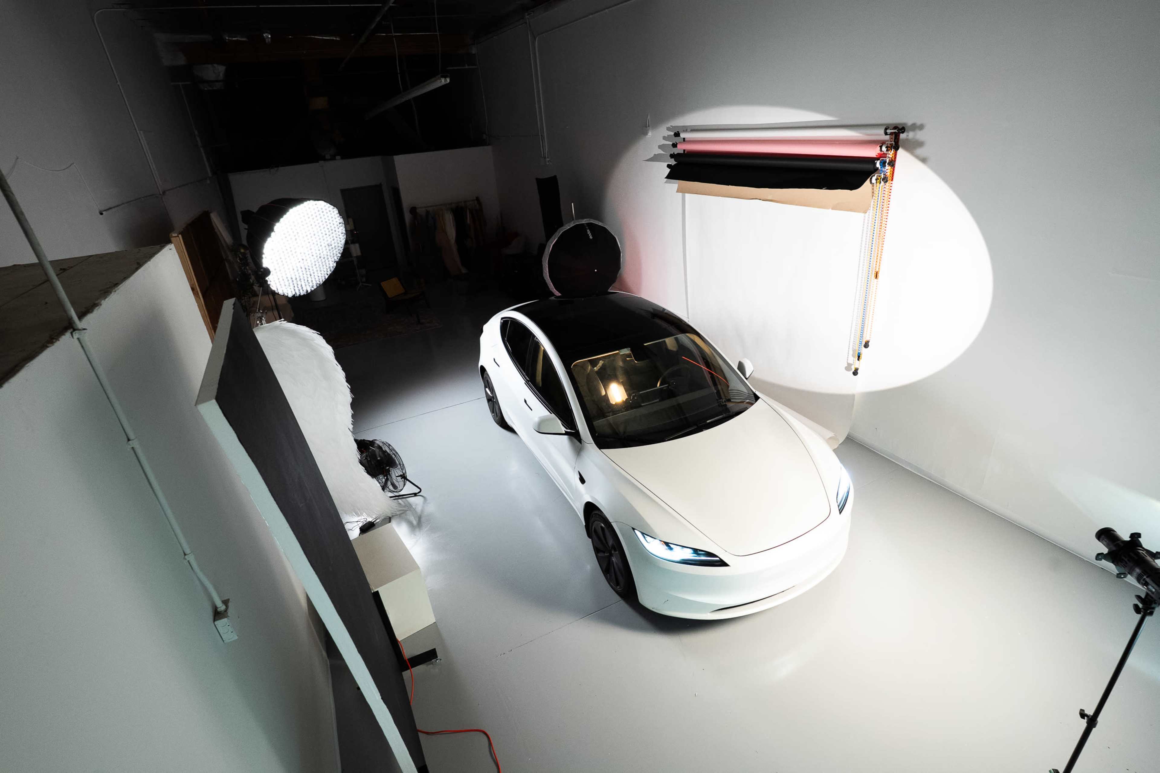 A white car is displayed in a well-lit photography studio with various lighting equipment and backdrops set up around it.