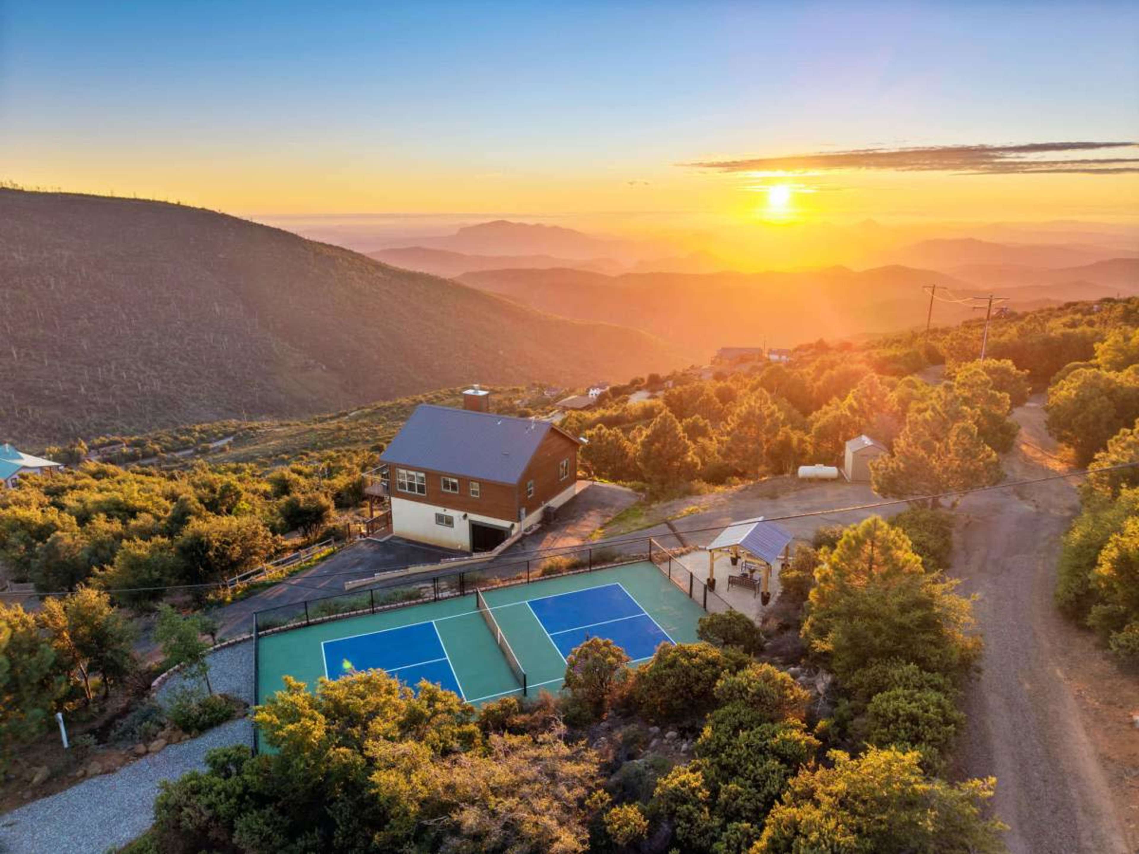 A hillside property with a house and two tennis courts overlooks a valley at sunset.