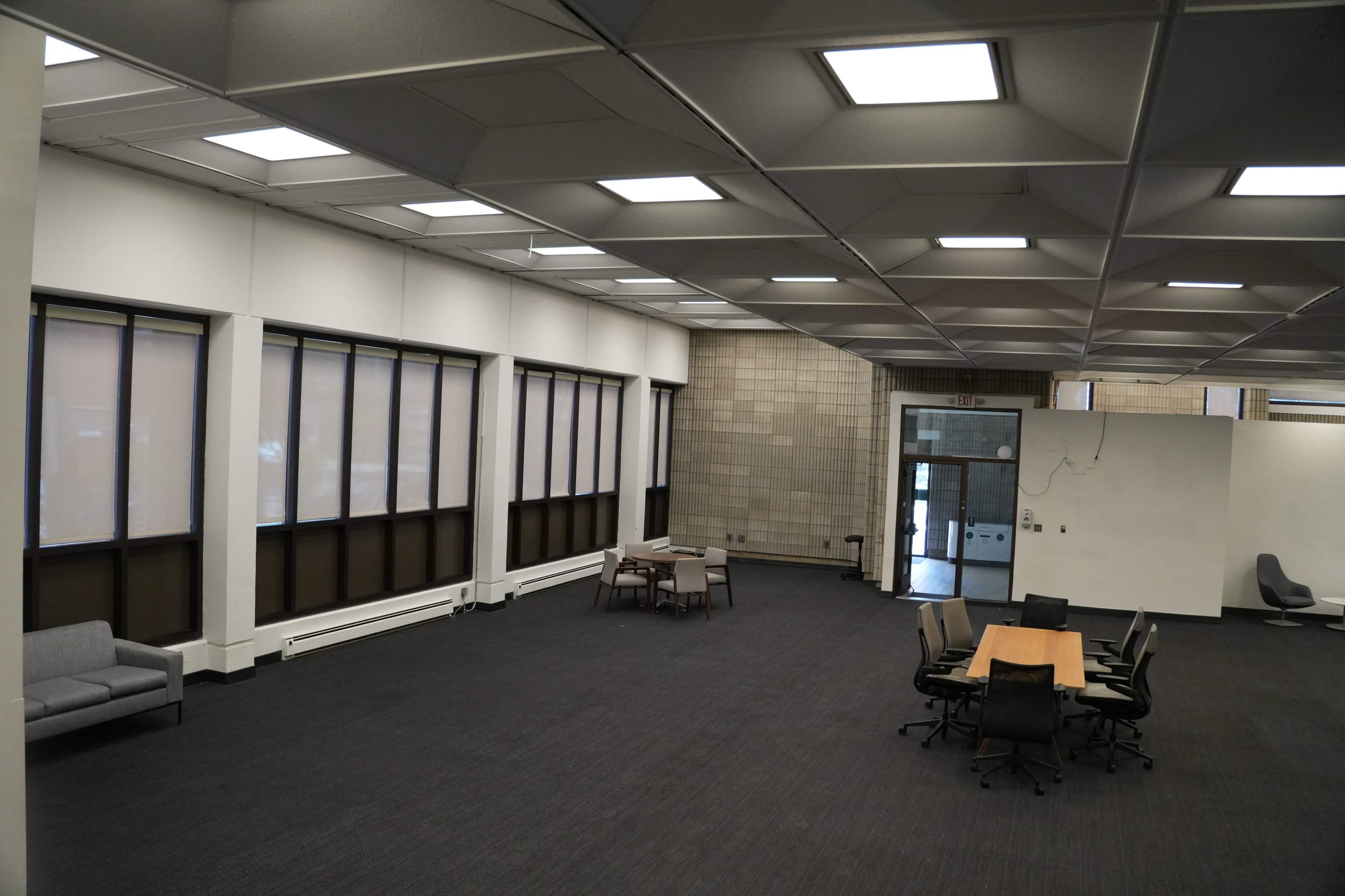 The image shows a spacious, empty office area with large windows, modern furnishings, and a central table surrounded by chairs.