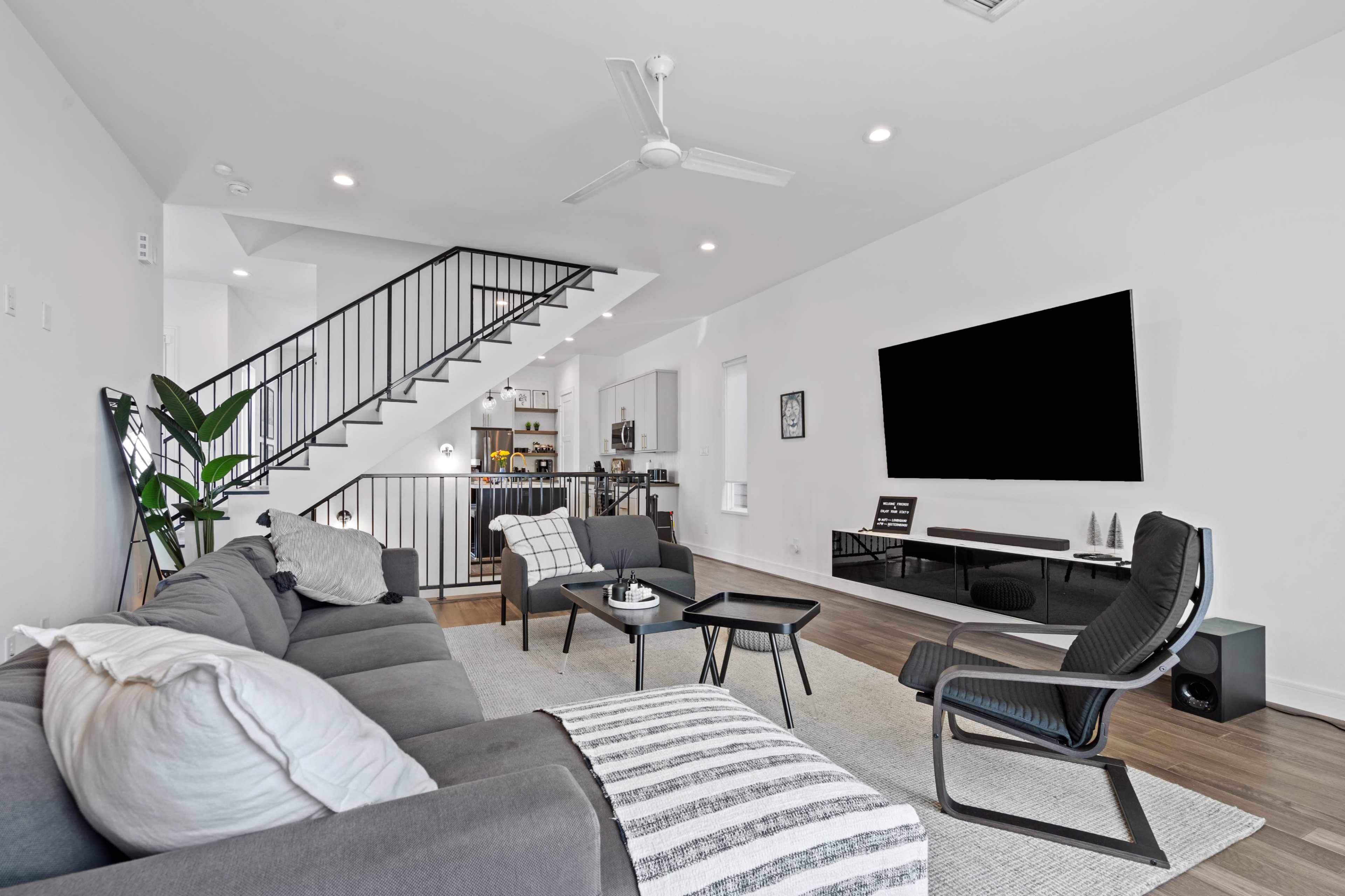 A modern living room features a gray sectional sofa, a striped area rug, and a large wall-mounted television, with a staircase visible in the background leading to a kitchen area.